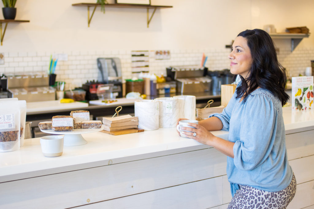 Dark-haired woman standing at a coffee counter holding a cup of coffee.
