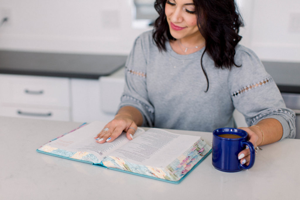 Dark-haired woman with her hand on an open Bible reading with a cup of coffee.