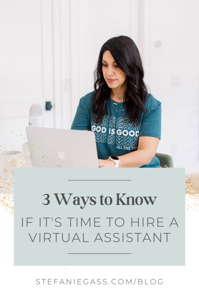 A long dark-haired woman is sitting at a desk with an open laptop in front of her. She is typing. She is wearing a blue graphic tee that says 'God is good all the time'. There is a podcast microphone next to her, as well as a coffee cup. The title of the graphic is: 3 ways to know if it's time to hire a virtual assistant. The link at the bottom of the graphic is stefaniegass.com/blog