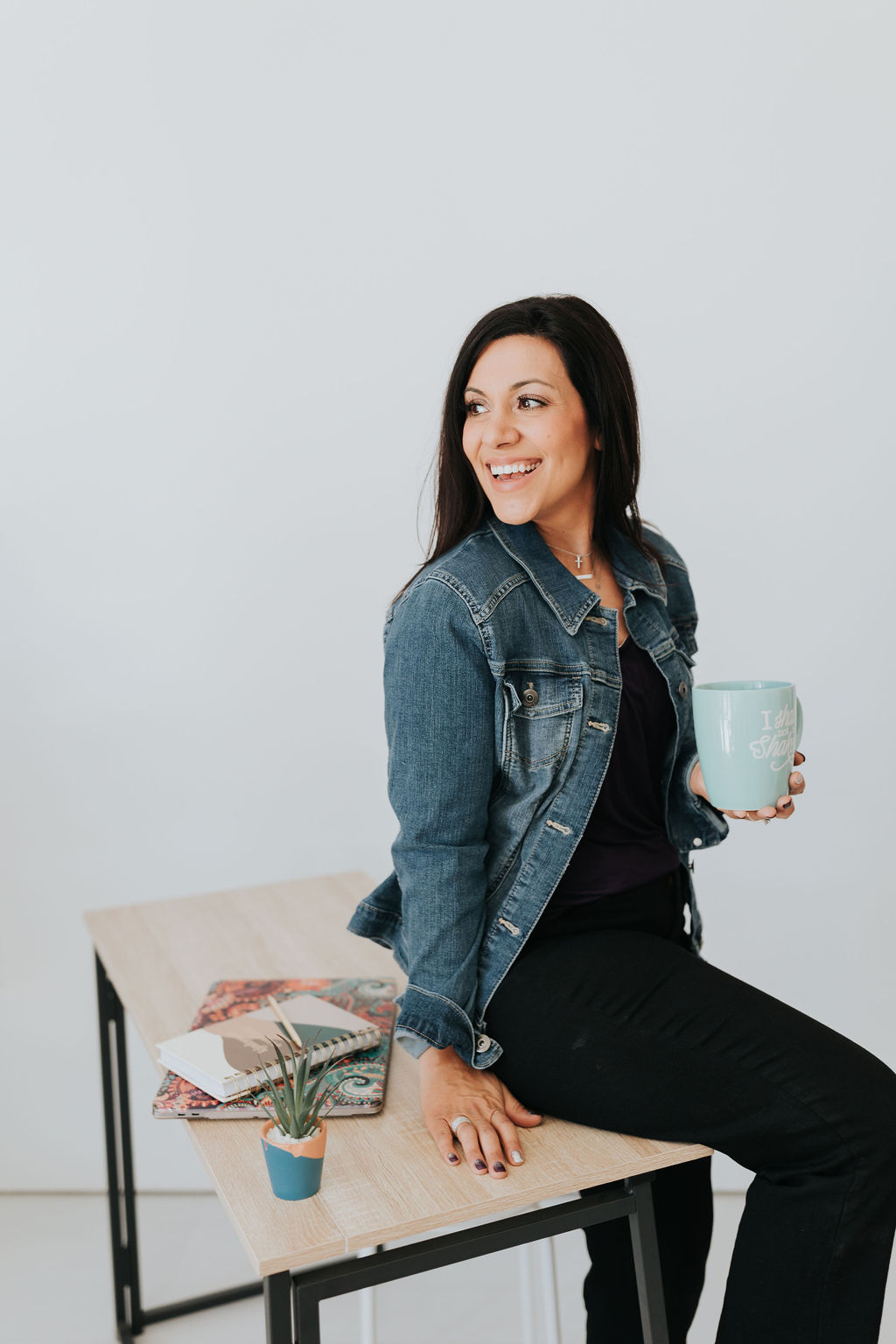 Dark haired woman sitting on her desk with a coffee mug. Stefanie Gas, online business coach.