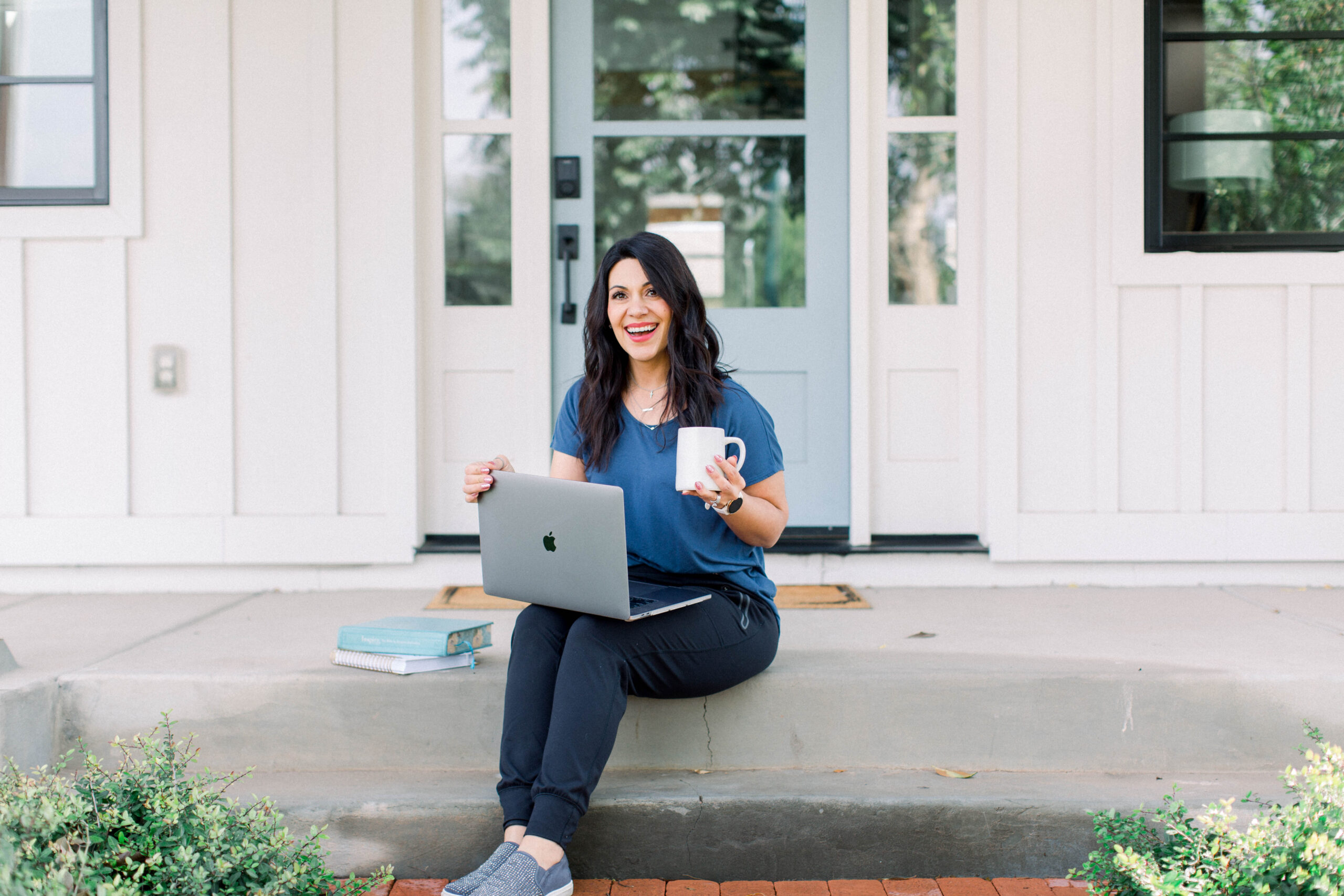Dark haired woman sitting on a step in front of her door, with her laptop and mug of coffee.