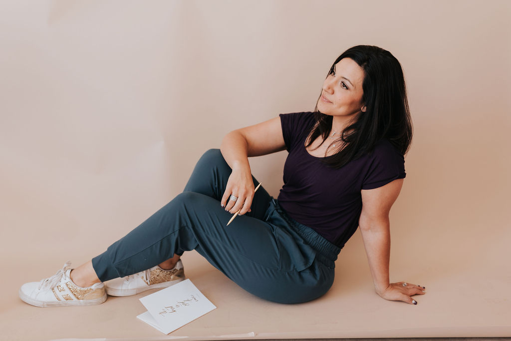 dark haired woman sitting on the floor, facing toward the left of the image. She looks comfortable, deep in thought. She is wearing a purple blouse and teal pants, resting on her left hand. Her right hand is holding a penical and there's a piece of paper on the ground near her.