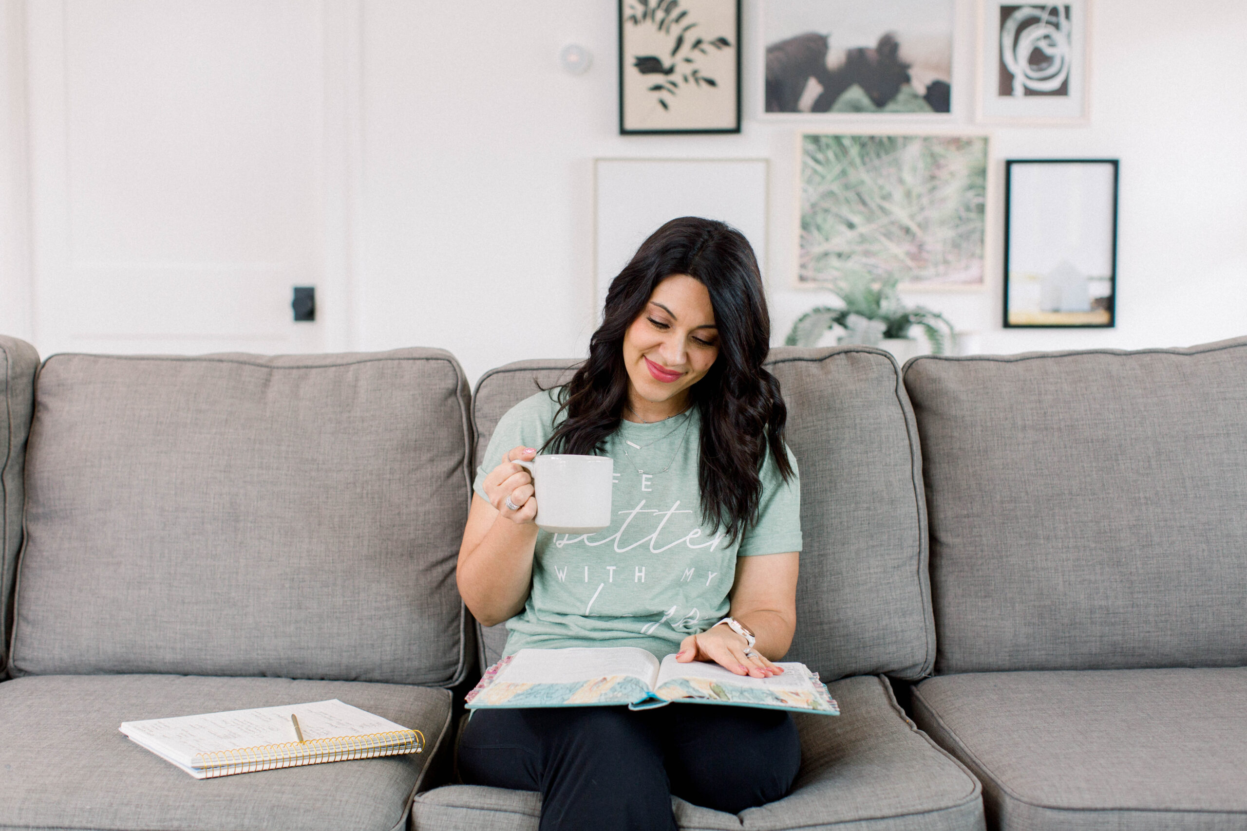 dark haired woman sitting on sofa reading a bible and drinking coffee