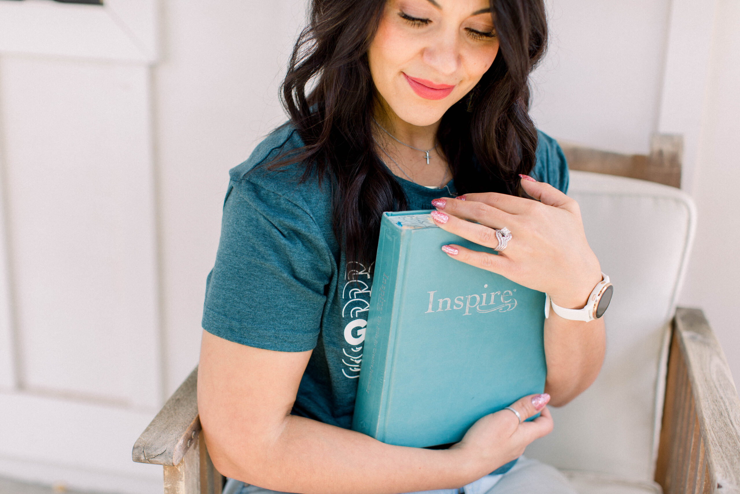 Dark haired woman in green t-shirt is sitting in a chair. She is looking down and holding a Bible.