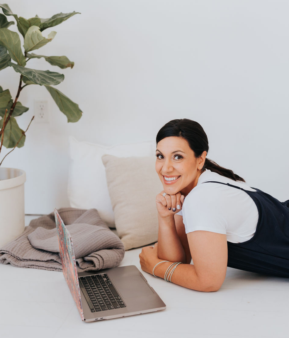 Dark haired lady leaning on her elbows smiling into the camera with a computer in front of her and two scatter cushions behind her.