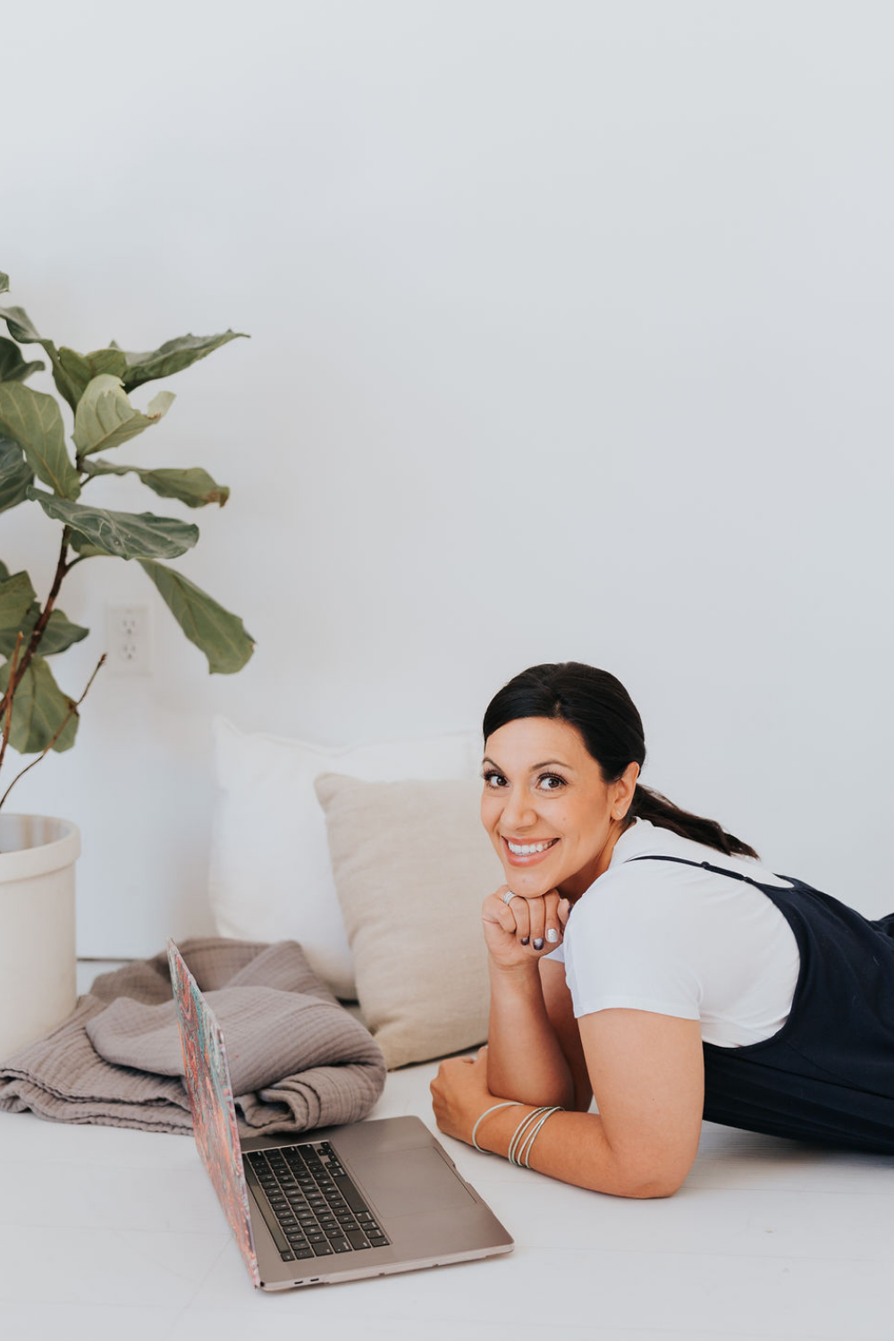 Dark haired lady leaning on her elbows smiling into the camera with a computer in front of her and two scatter cushions behind her.
