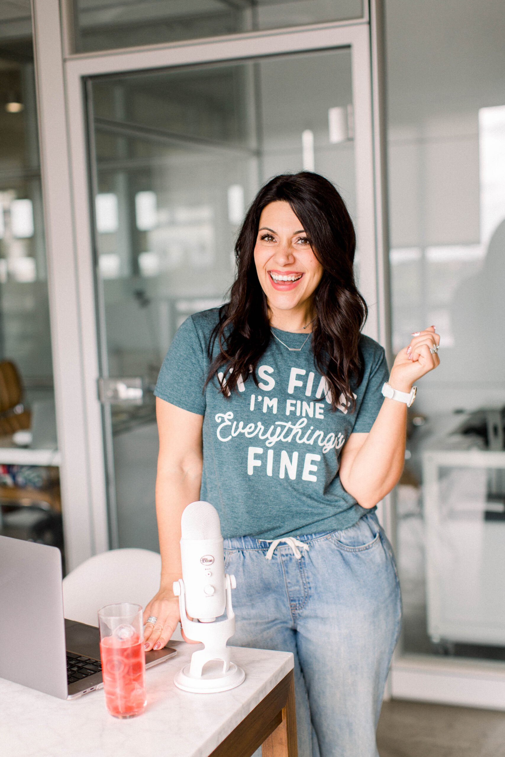 dark haired woman in a green shirt and blue jeans, is standing in front of a glass wall with a table in front of her. on the table is a laptop, white microphone, and a glass of juice. She is smiling at the camera.