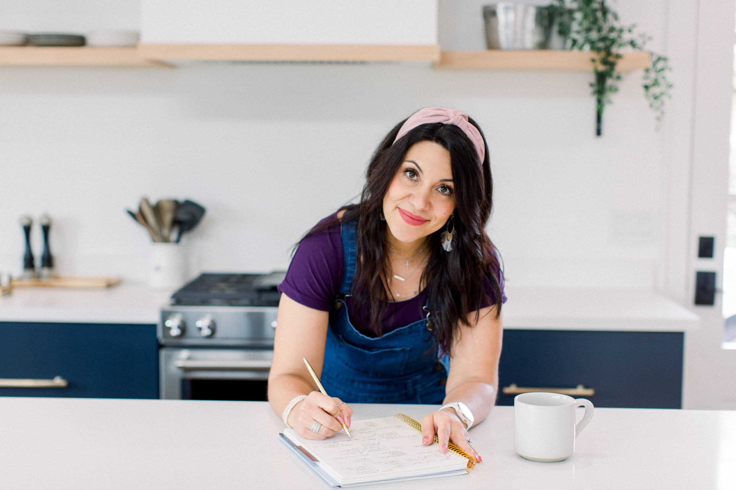 dark haired woman leaning on a kitchen counter with a notebook, writing a list. She is wearing a pink headband, purple blouse, and denim overals. She is smiling at the camera and there's a white coffee mug next to her left hand.