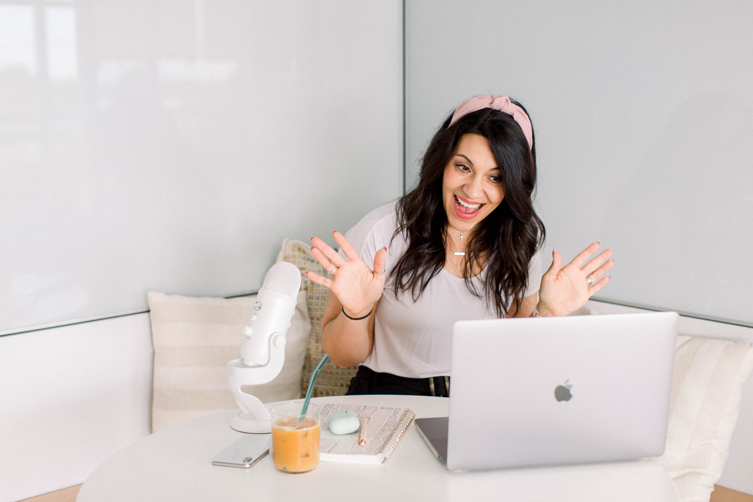 dark haired woman with a pink headband sitting at a table. She is in front of a laptop, smiling toward the screen, smiling and has both hands palms forward, showing all ten fingers. She has a notebook and pen next to the laptopn with a glass of juice and a white microphone.