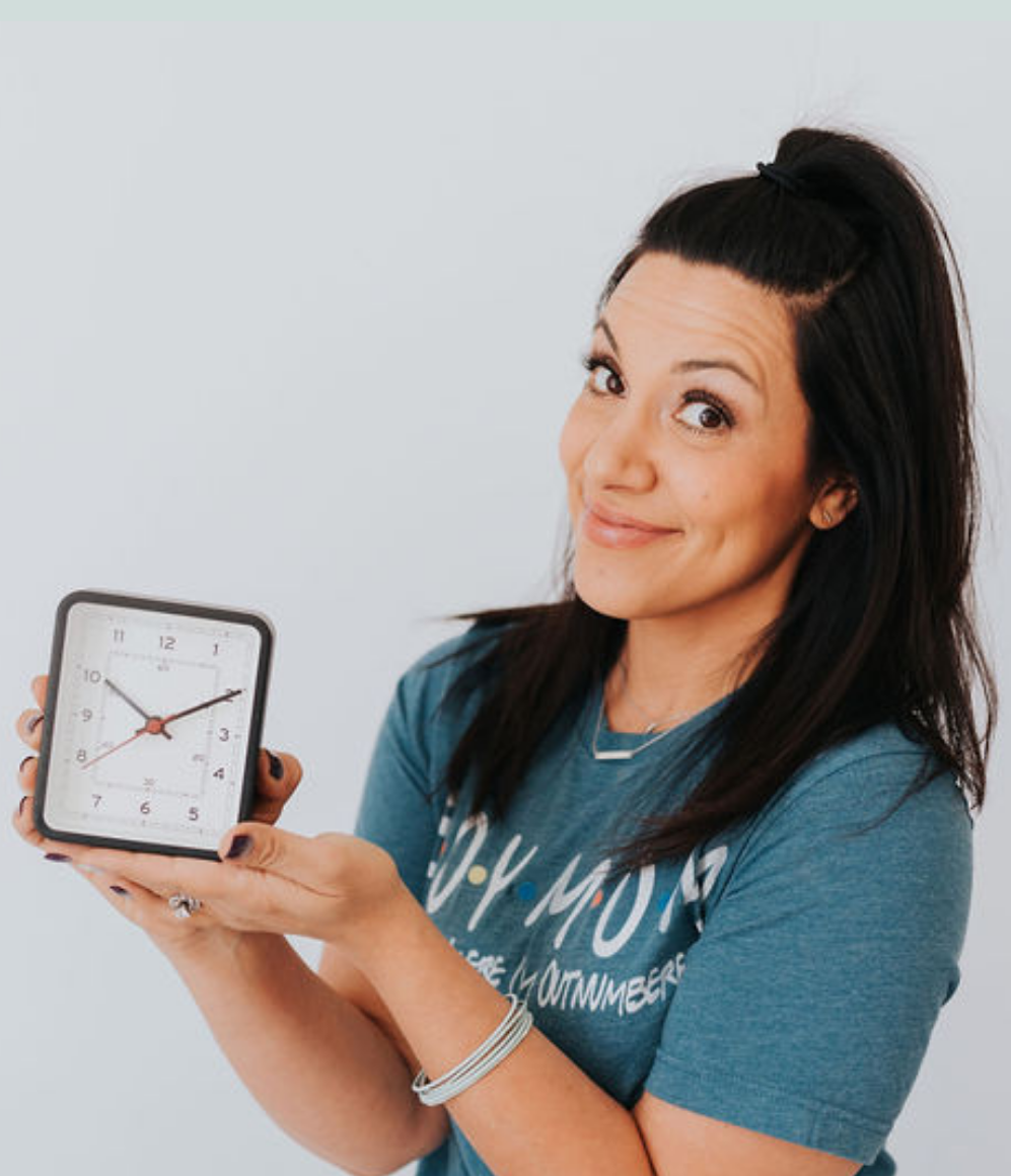 dark haired lady wearing a green t shirt and pants and holding up a clock in both hands to the right of her body, smiling into the camera