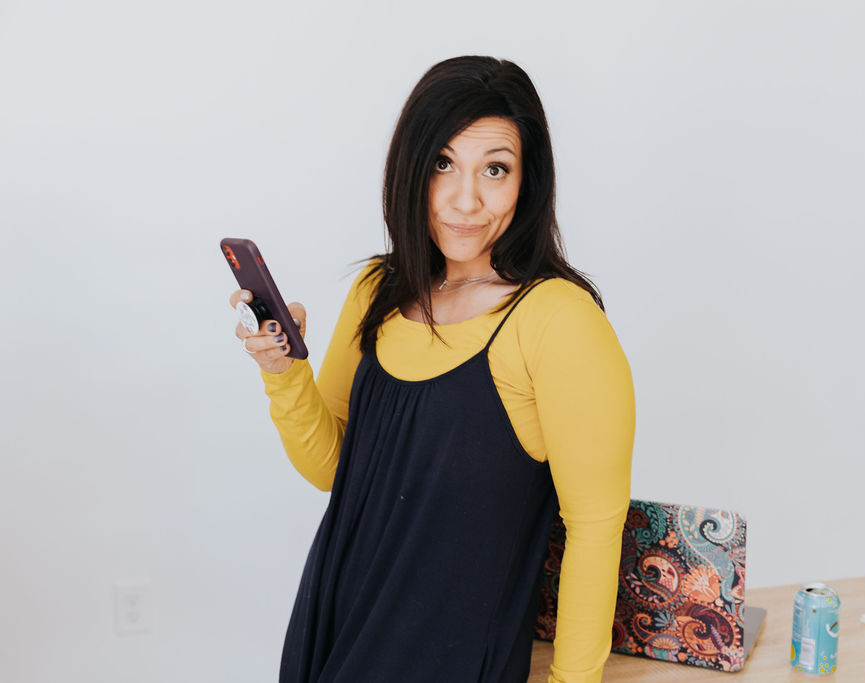 dark haired woman wearing a yellow blouse under a black tank dress. She is leaning against a desk holding a cell phone. She has a scowl on her face. There is a can of fizzy water and a laptop on the desk.
