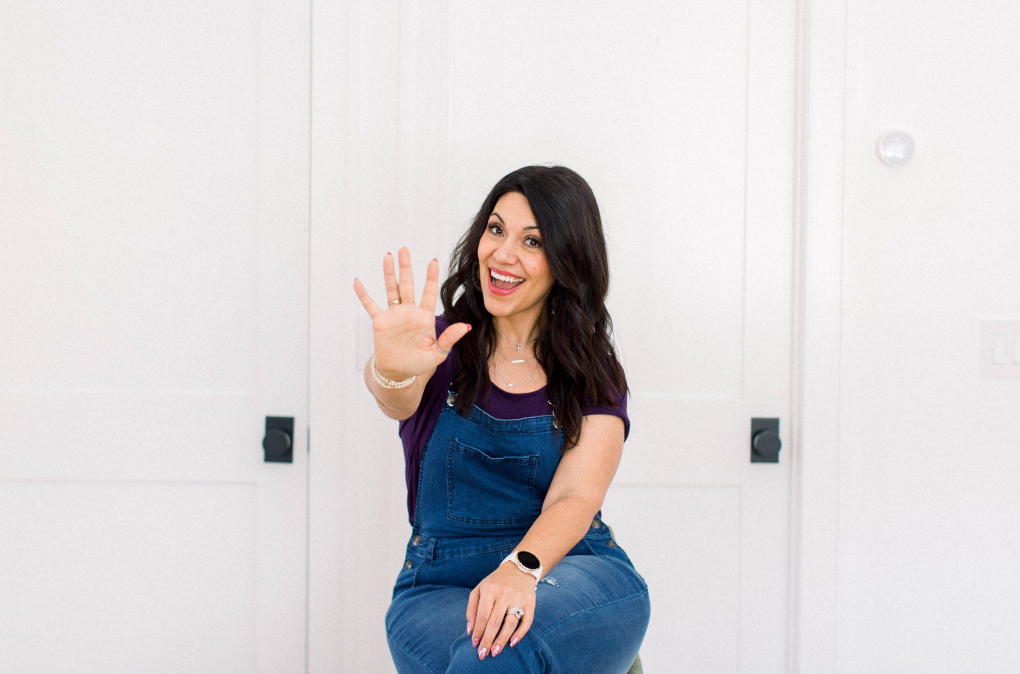 dark haired woman sittin on a stool in front of a white door. She is wearing a purple shirt with denim overalls. Her right hand is extended with all five fingers showing indicating a quantity of five.