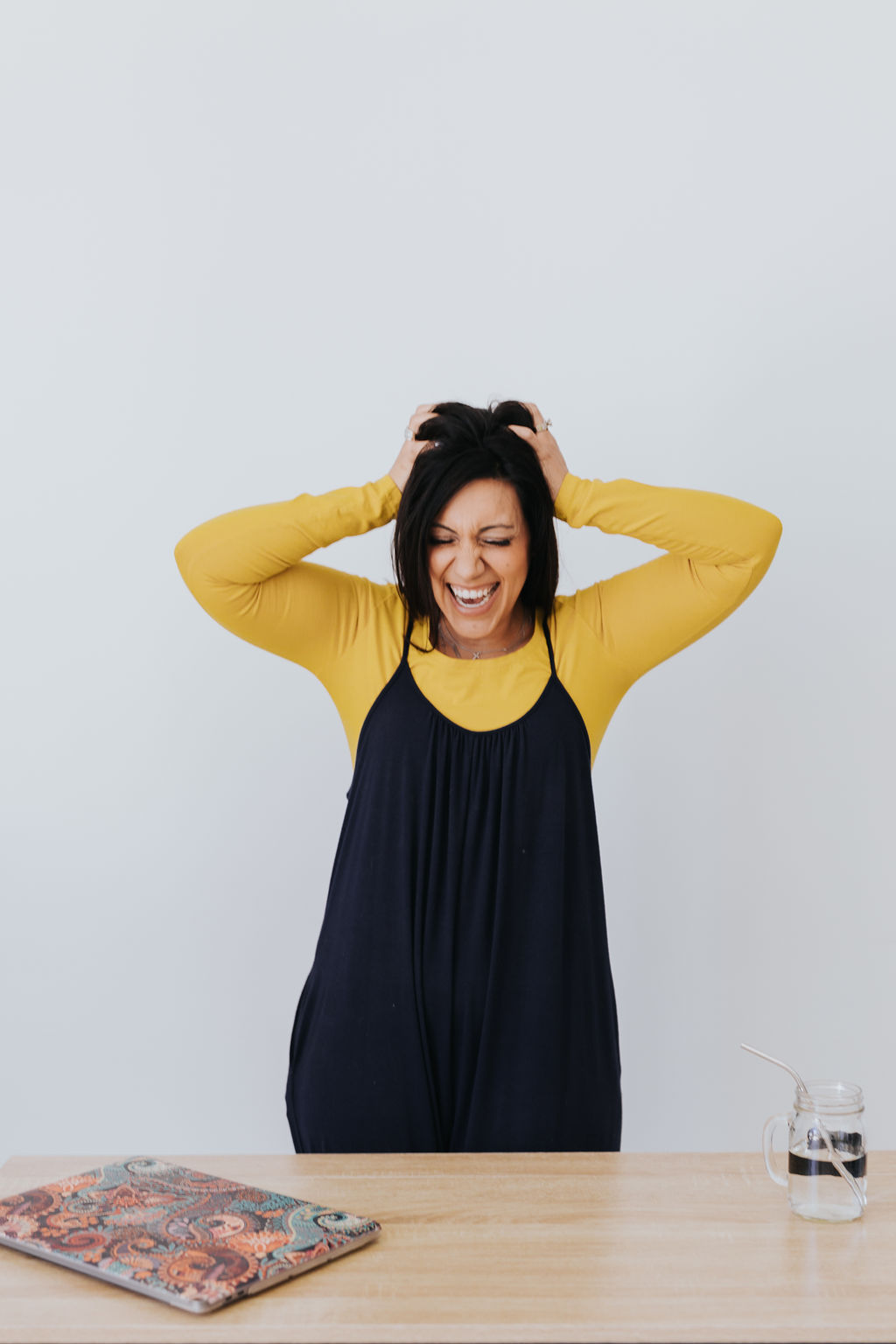 dark haired woman in a black jumper dress with a yellow long-sleeved blouse. She has her hands mussing up her hair and there is a scatterbrained look on her face.