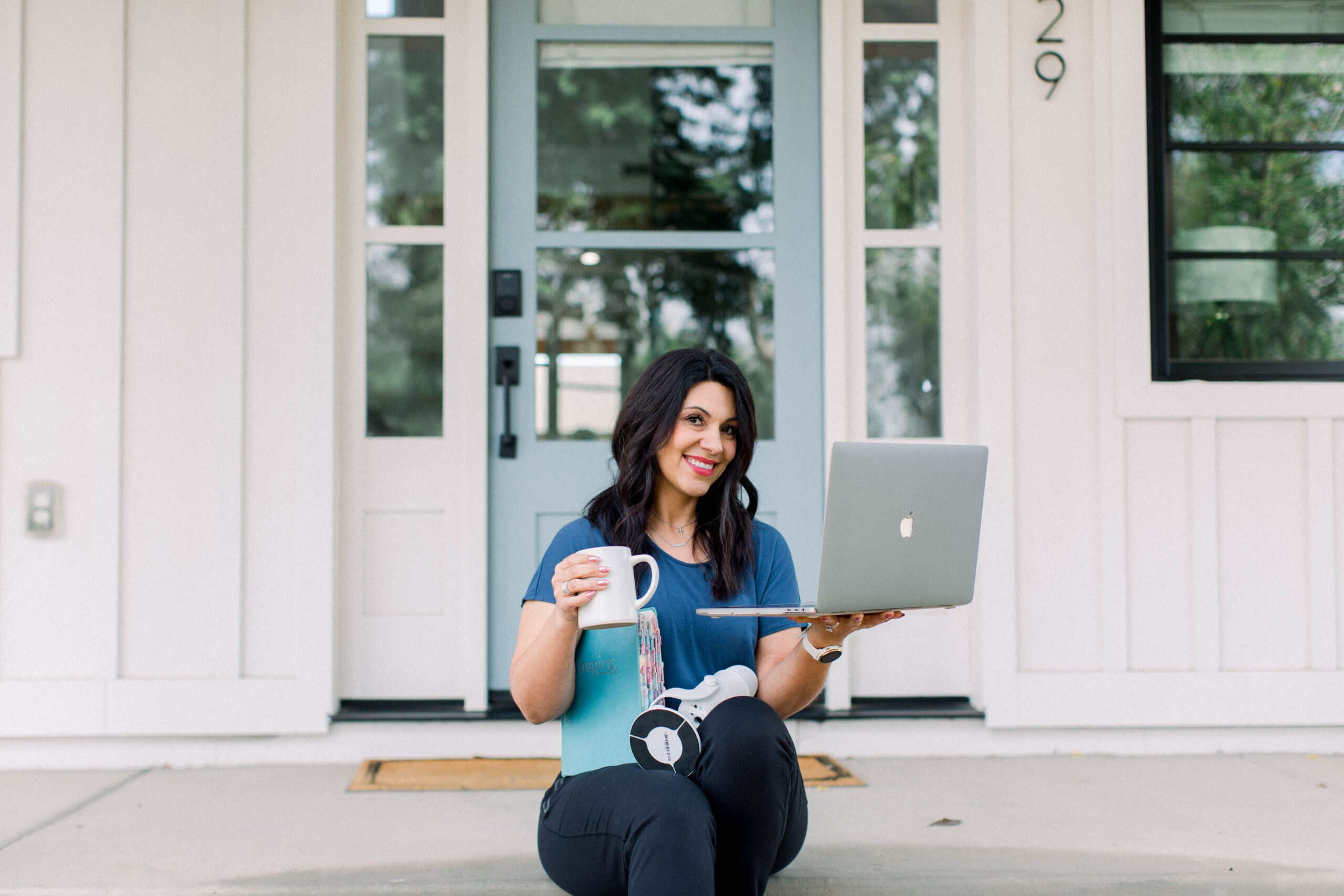 photo of a dark haired woman sitting on the steps in front of a house with her laptop, bible, coffee mug, and microphone in her lap.