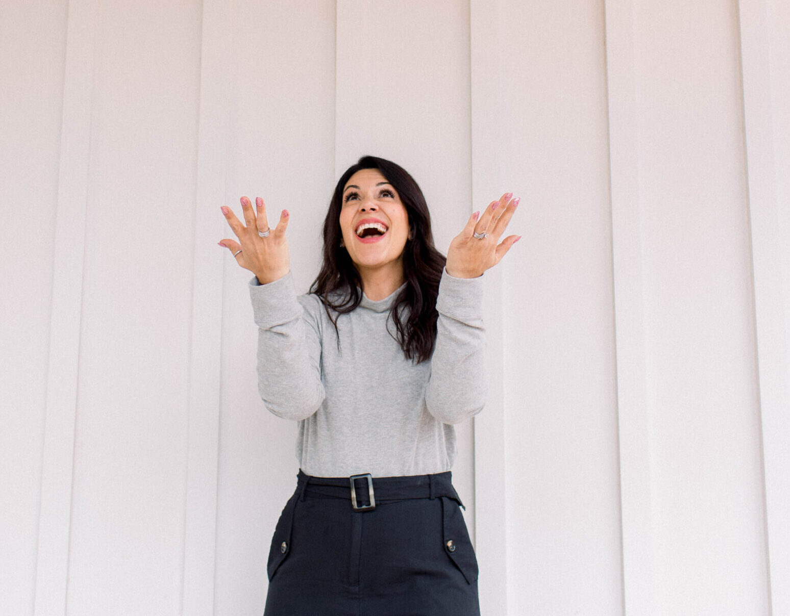 dark haired woman with her back against a white wall is wearing a grey sweater and black pants. her face is looking up and she is smiling. Her arms are bent at the elbows with her palms facing up.