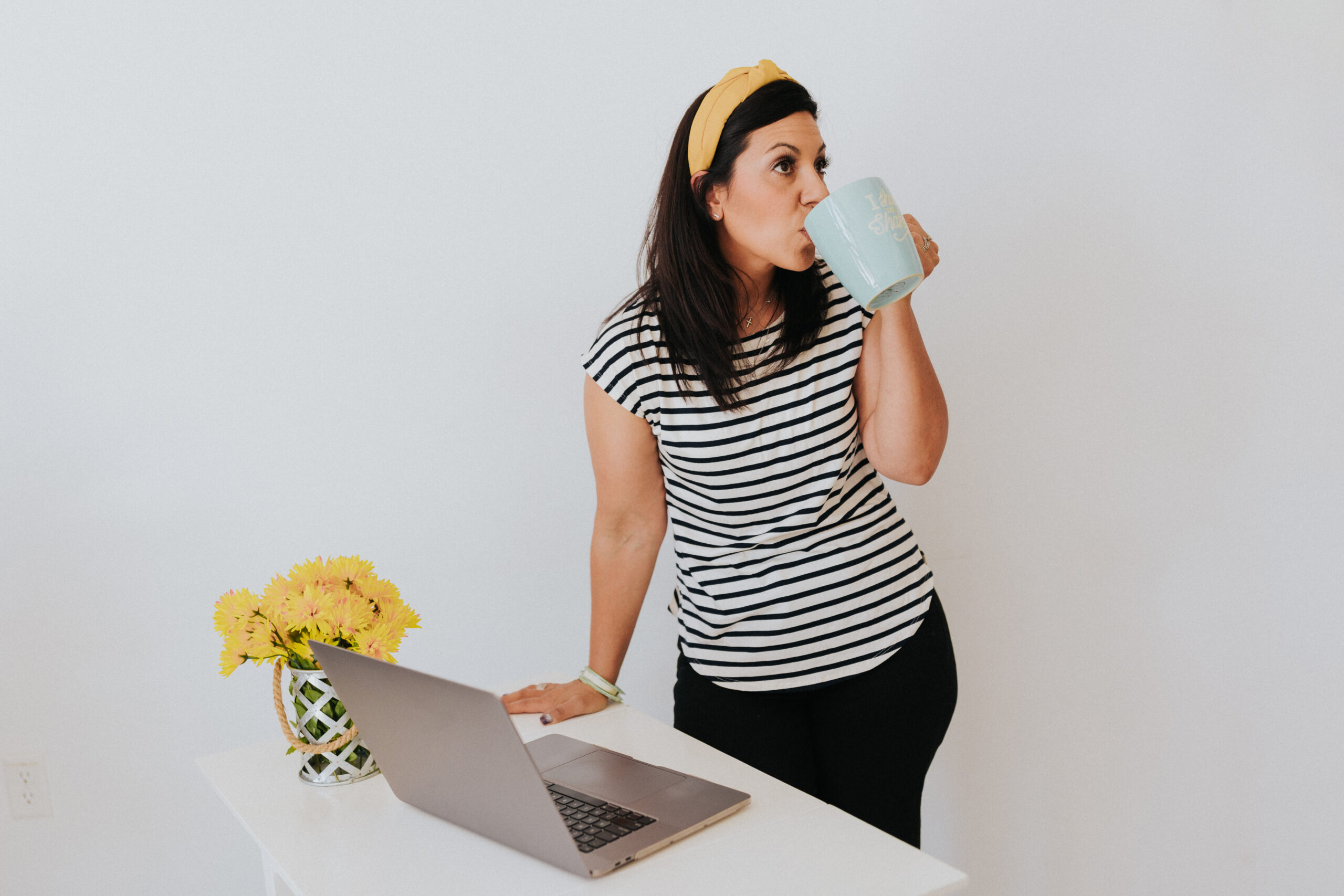 photo of a dark haired woman standing beside a desk with a coffee mug in her hand and laptop on the table