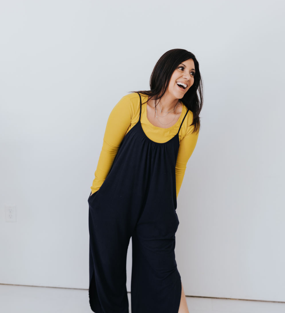 Dark haired lady in yellow top and black jump suit with hands in her pockets leaning towards the left of the picture, smiling at someone out of the picture