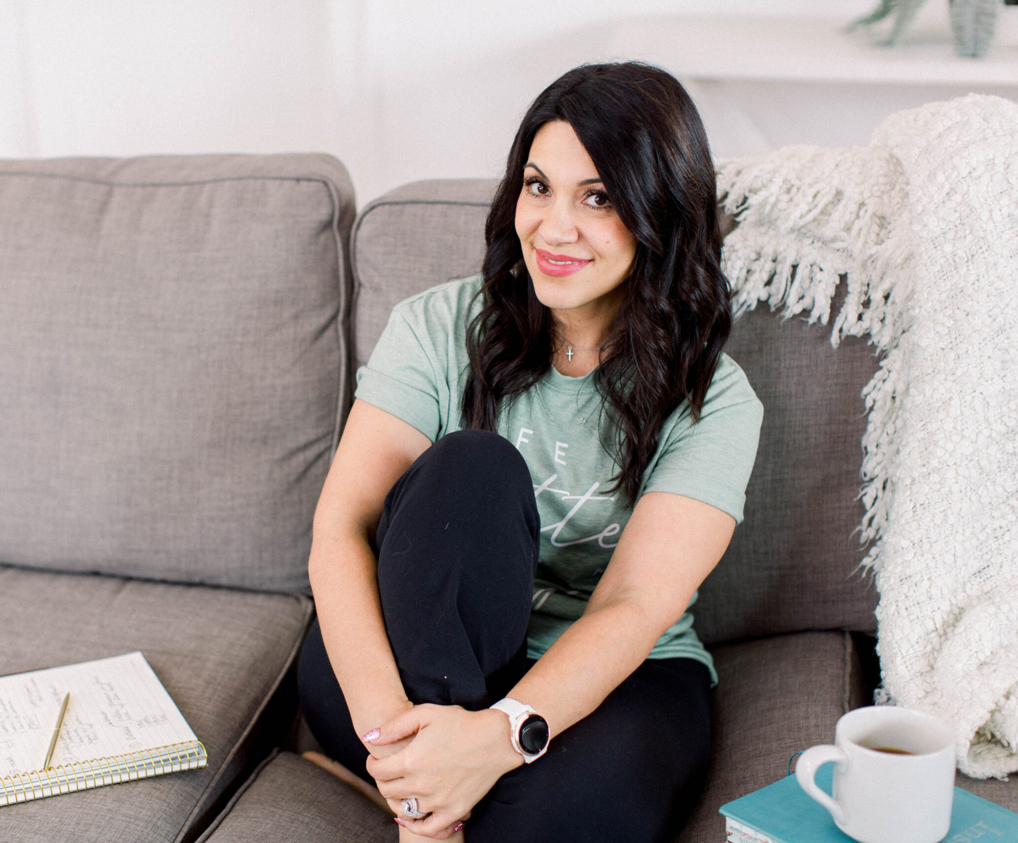 dark haired woman sitting on a couch with her right knoee drawn up to her chest. She is wearin a light green t-shirt and black slacks. Next to her is a notebook and pen on one side and a cup of coffee on the other. She is smiling at the camera.