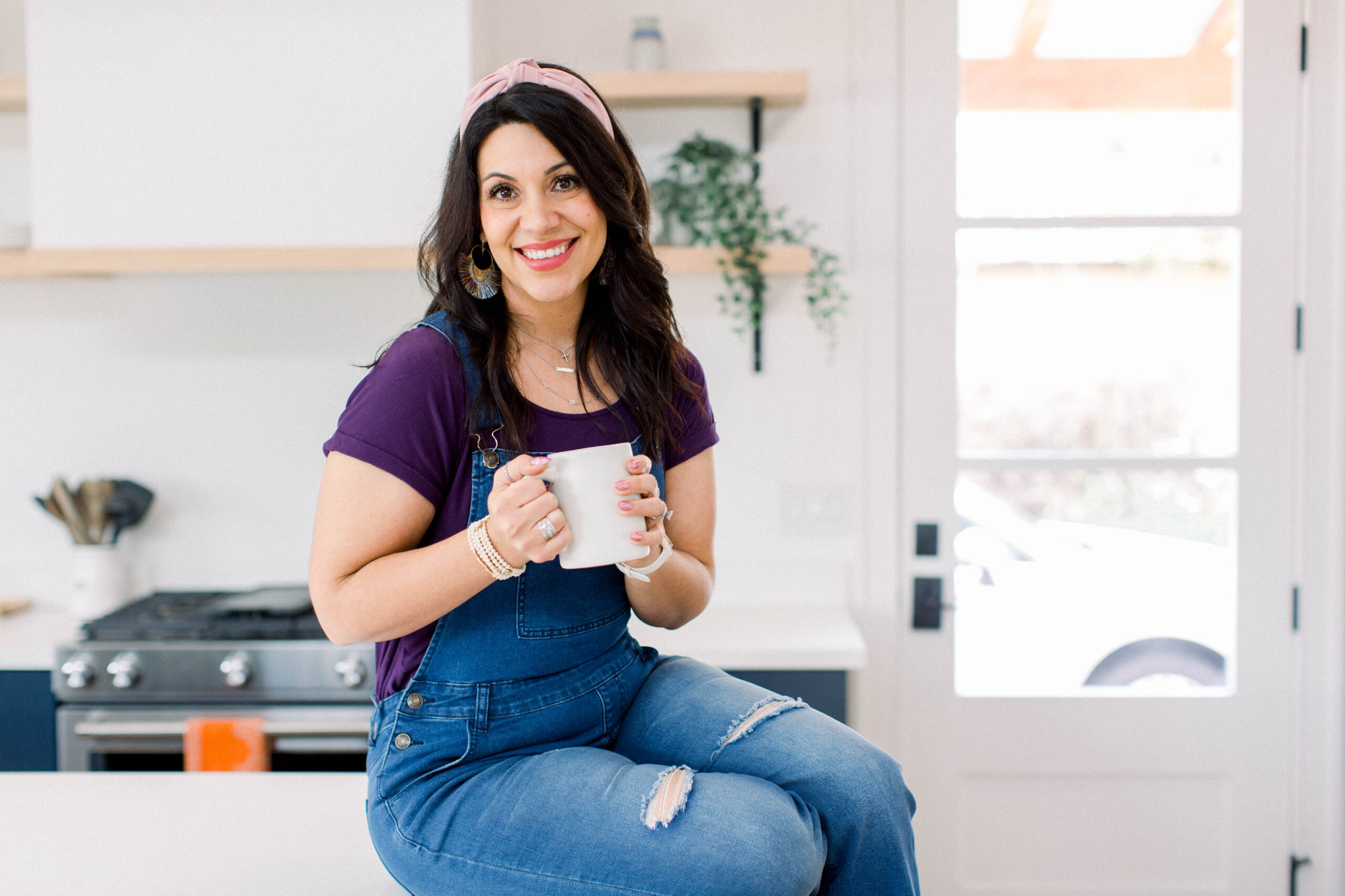 dark haired woman sitting on a kitchen counter. She is wearing denim overalls, a purple t-shirt, and a pink headband. She is smiling at the camera and holding a white mug.