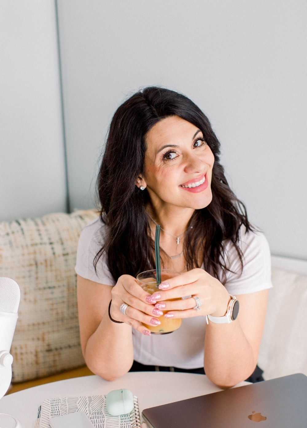 dark haired lady sitting on a sofa at a table with a closed laptop and phone, airpods, a notebook and a microphone. She is smiling into the camera with a glass of orange juice held in both hands