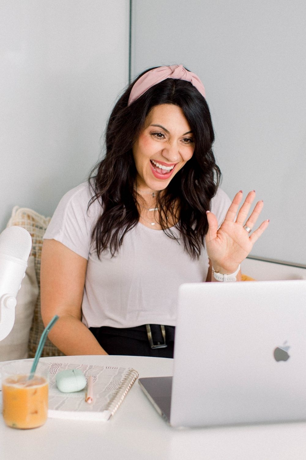 dark haired lady sitting on a sofa with a pink head back and a white t shirt. She is smiling and waving into a laptop on a table in front of her. There is also a glass of orange juice with a straw in it on the table.