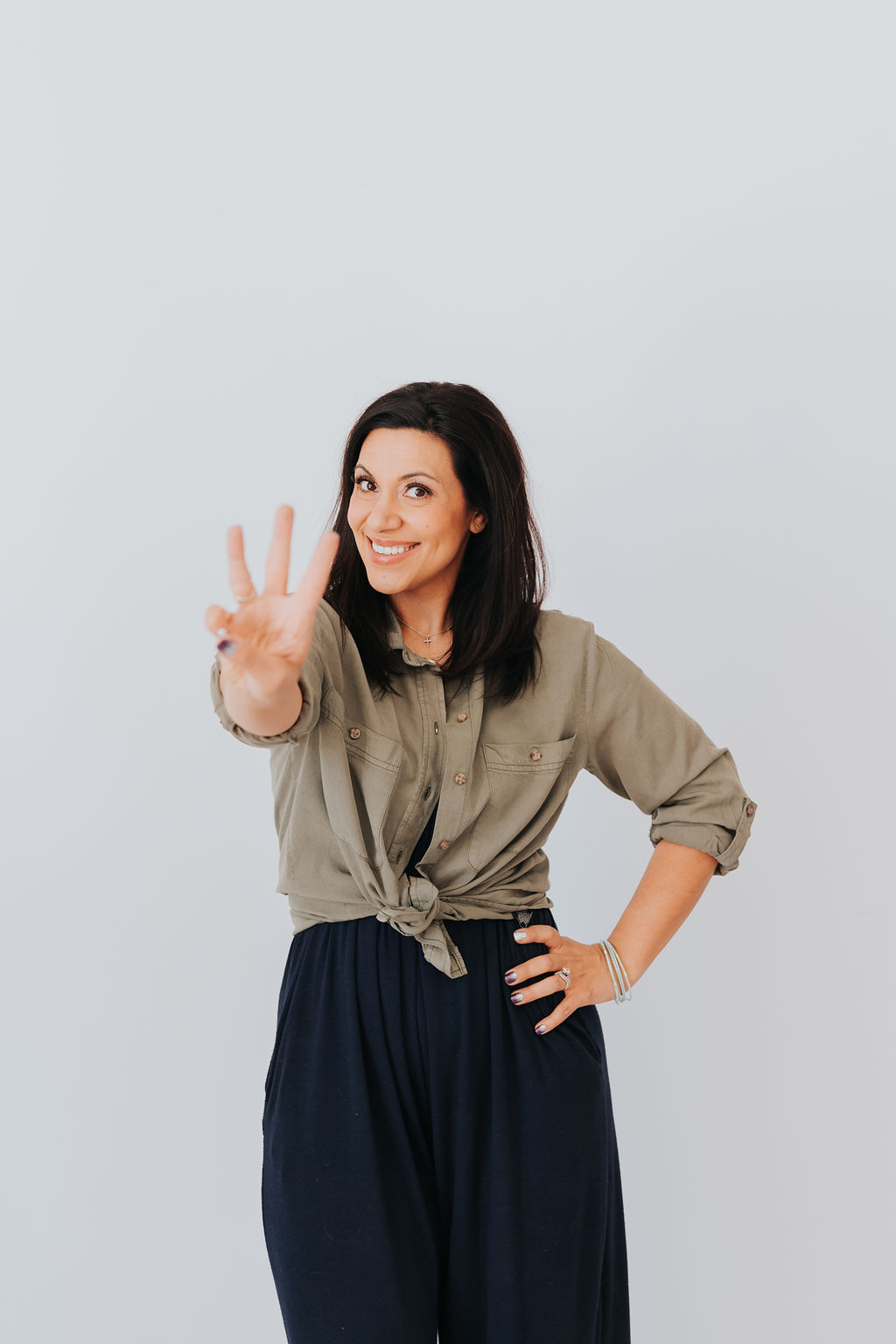 dark haired woman with a khaki blouse and black skirt is standing, facing the camera. She has her left hand on her hip and her right hand is raised up near her shoulder while holding up three fingers. She is smiling.