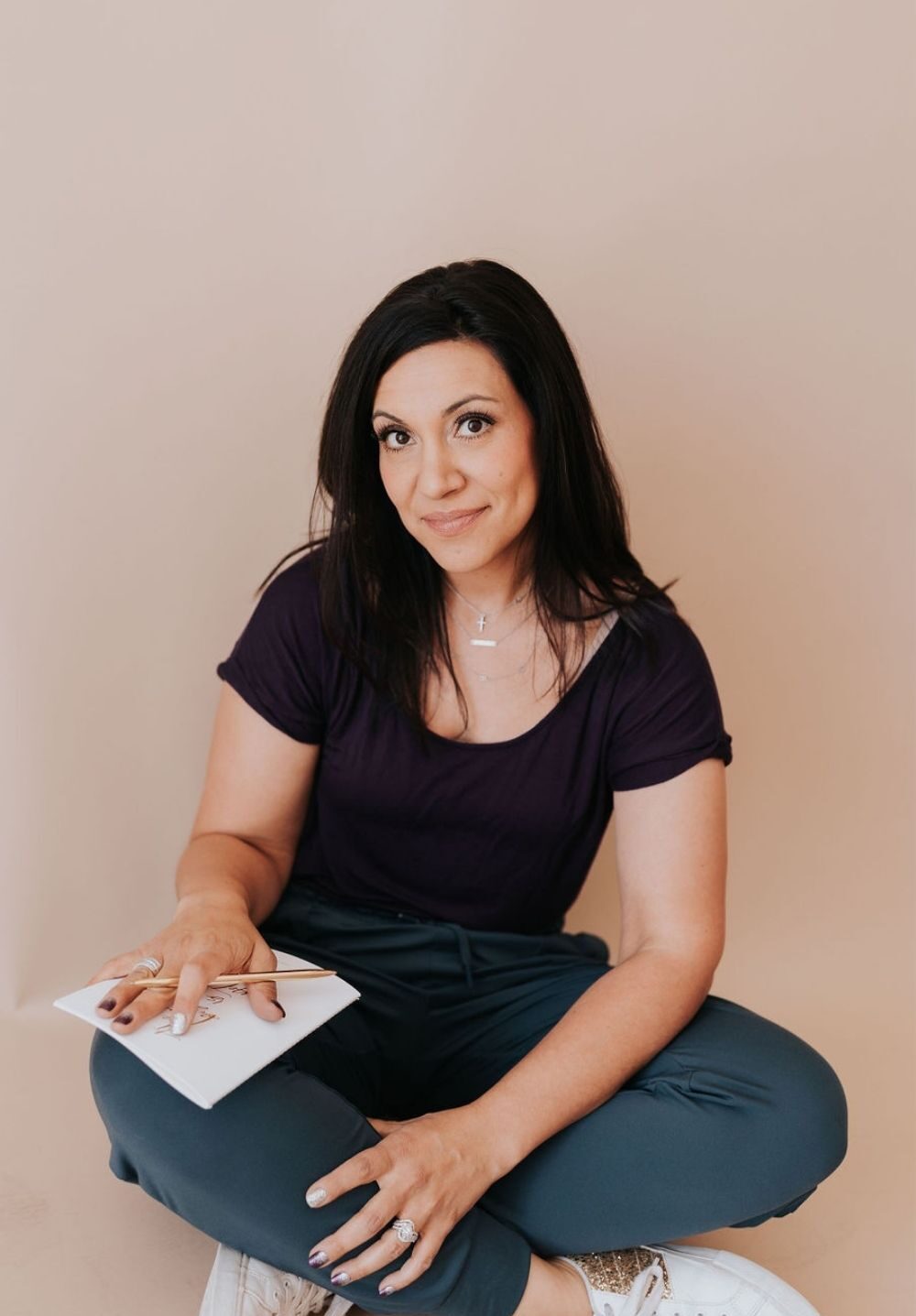 dark haired lady sitting cross legged looking into the camera with a book on her lap and a pen in her right hand, left hand resting on her foot.