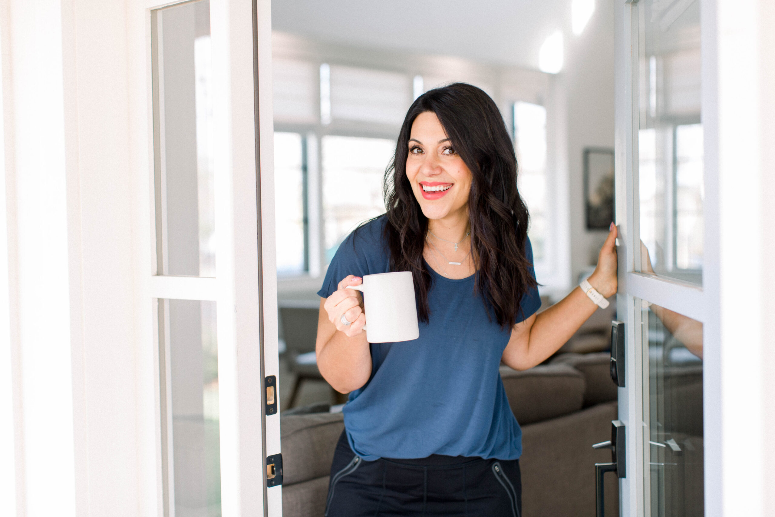 dark haired woman standing in a doorway. She has her left hand on the edge of the door and is holding a white mug in her right hand. She is wearing a blue blouse and black jeans. She is smiling at the camera.