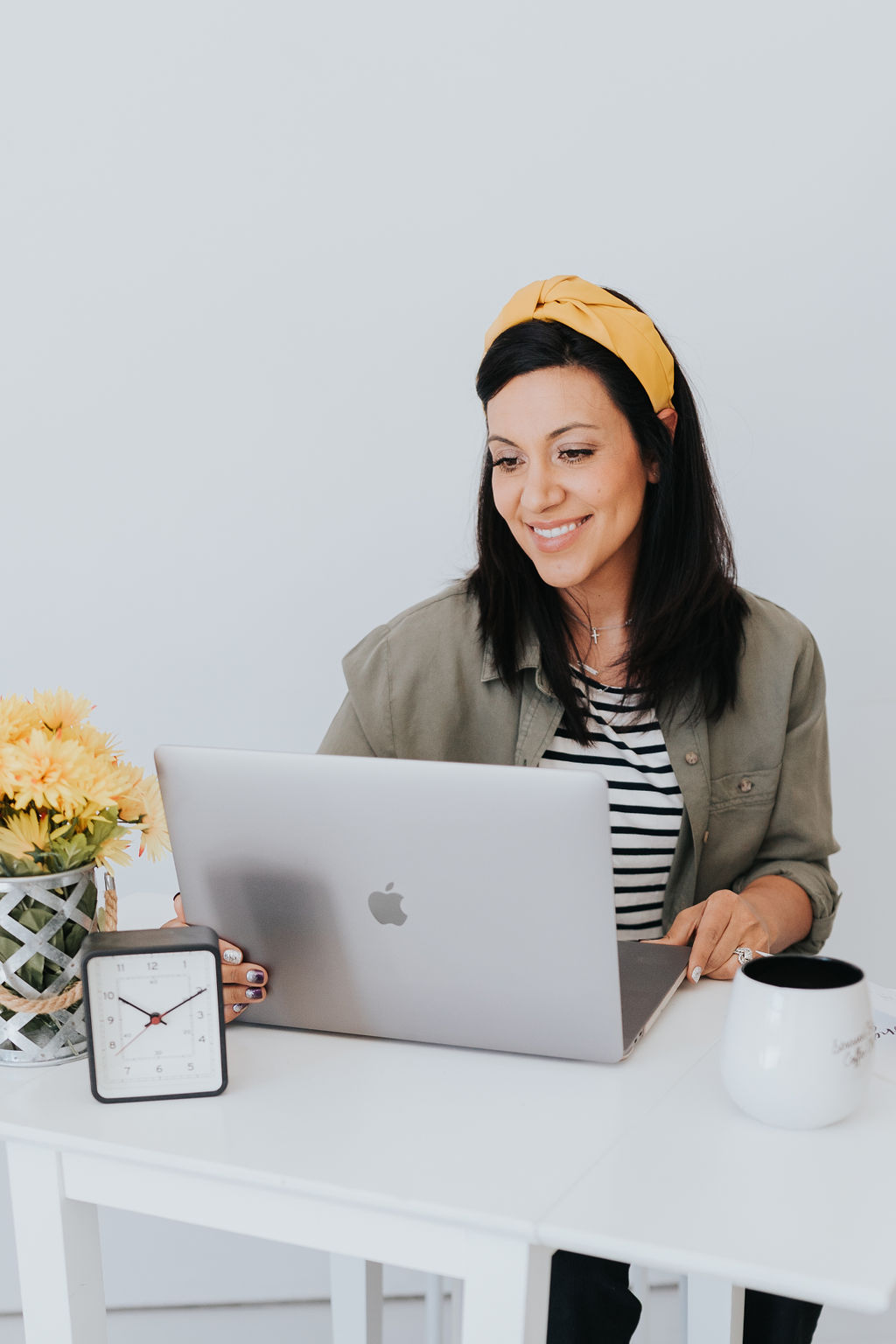 dark haired woman sitting at a desk with her laptop open in front of her. Also on the desk is a vase of yellow flowers, and alarm clock, and a mug of coffee. She is wearing a yellow headband and a black and white striped shirt.