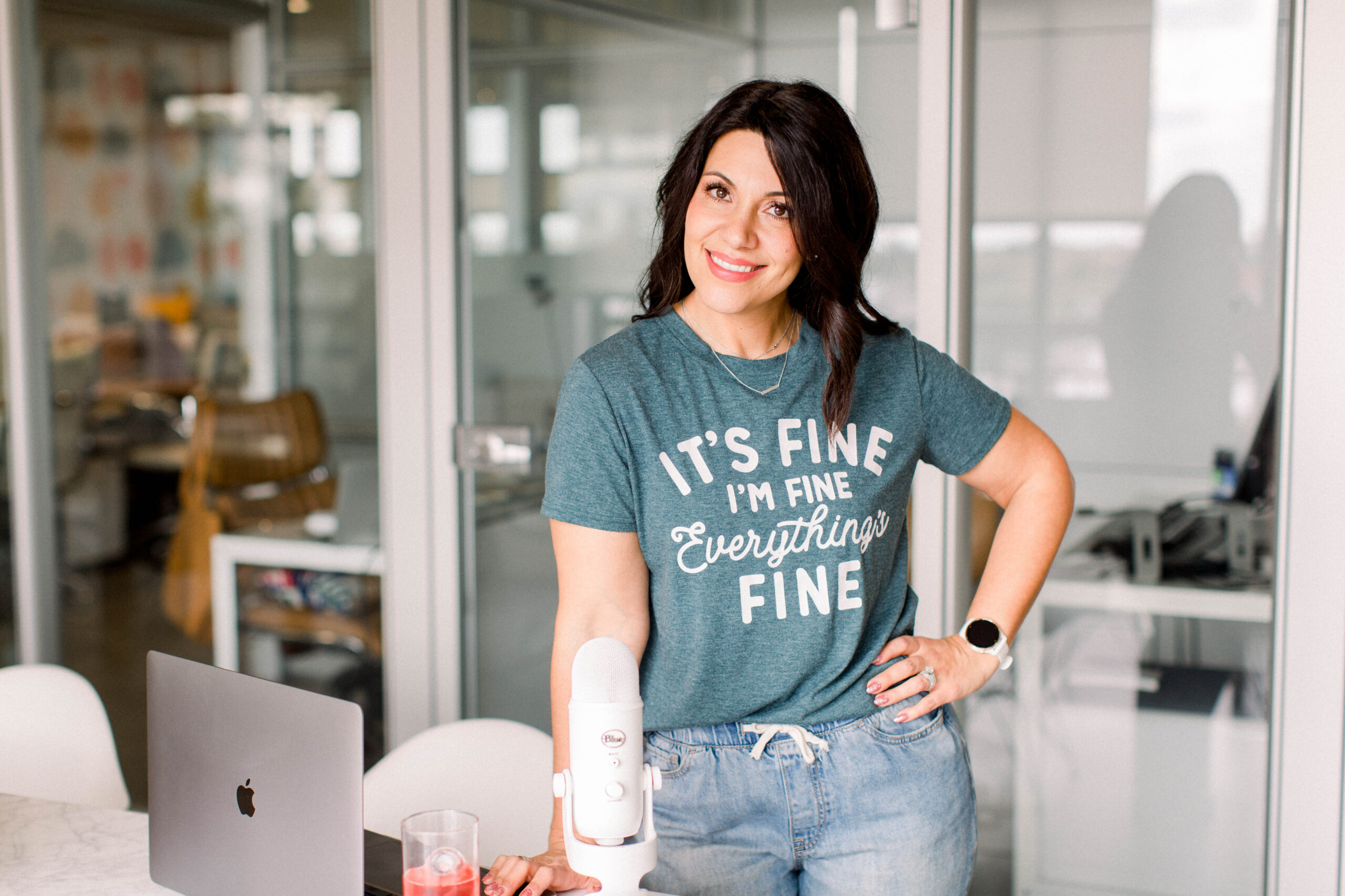 dark haired woman standing in front of a glass wall with a table in front of her. She is wearing a green t-shirt and jeans. There is an open laptopn and white microhpone on the table. Her left hand is on her hip and she is smiling toward the camera.