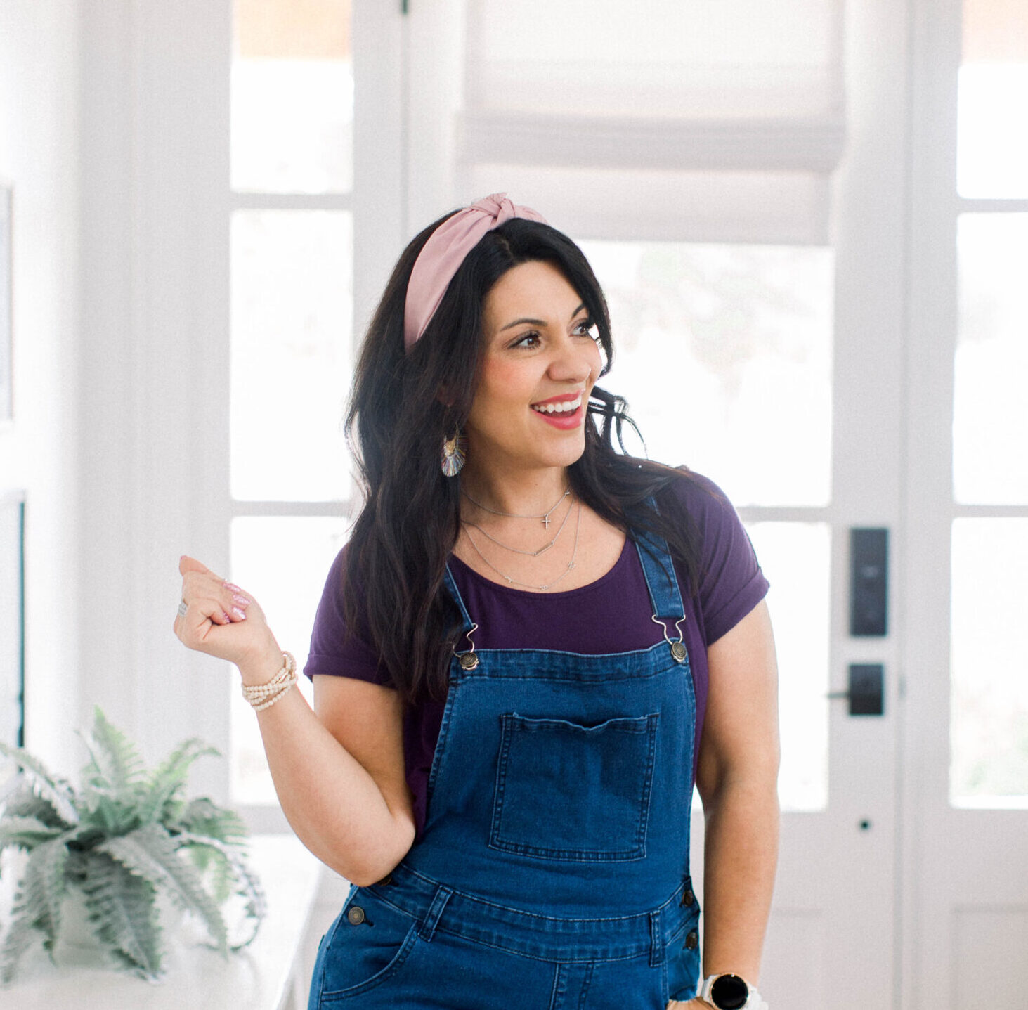 dark haired woman wearing denim overalls and a purple shirt, has her left hand resting at her pocket and her right hand up in the finger-snapping position. She is standing in a mostly white room with a plant on a shelf behind her.