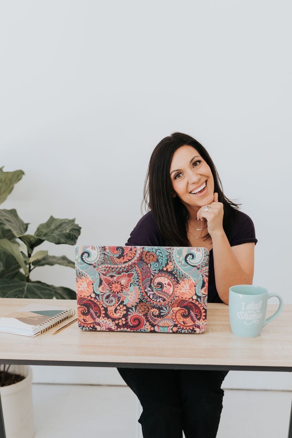dark haired lady sitting at a desk with her chin resting on her left hand smiling into the camera. To her right is a house plant and on the desk in front of her is a laptop, a coffee cup and a notebook and pen.