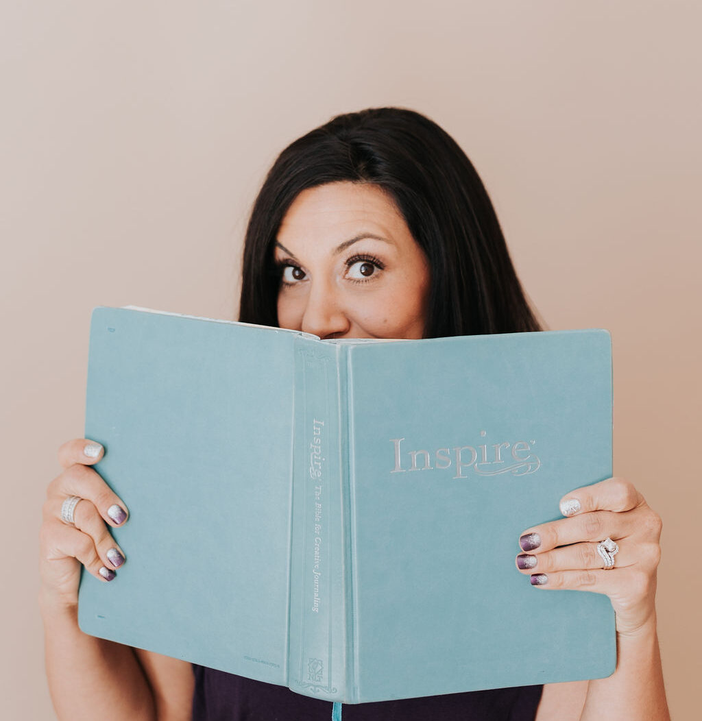 dark haired woman holding an open blue Bible in front of her face. You can see her eyes above the top edge.