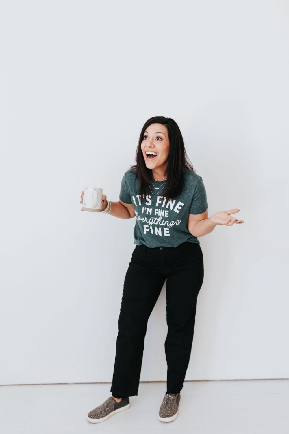 dark haired lady wearing black pants and a green t-shirt holding a mug in her right hand with her left hand up to her side, palm up smiling at someone off the right of the camera