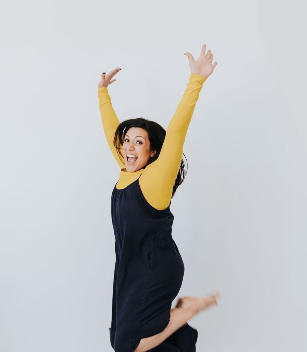 dark haired lady wearing a mustard top and a black jump suit jumping in the air with her hands above her head and smiling into the camera.