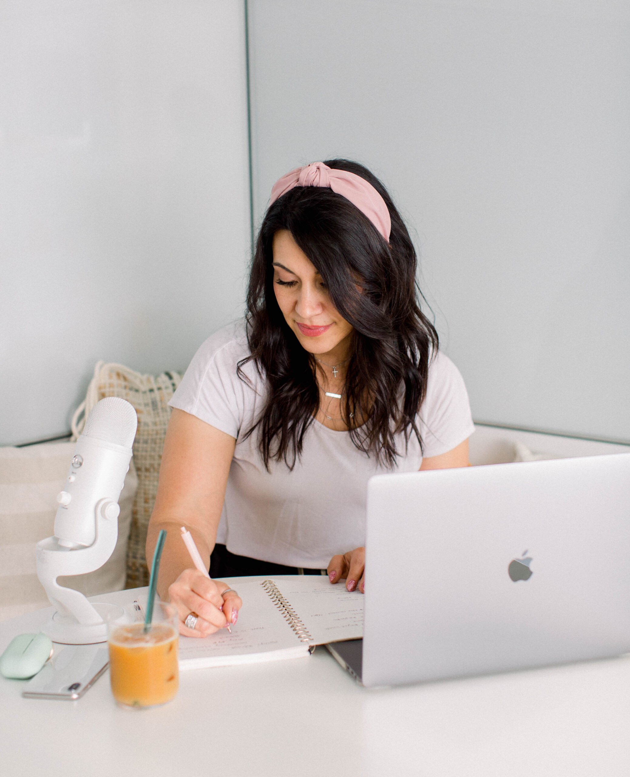 dark haired woman wearing a pink headband and a white tshirt sits at a desk. There is an open laptop on the desk, and she is writing in a journal. There is an iced coffee and podcast mic on the desk.