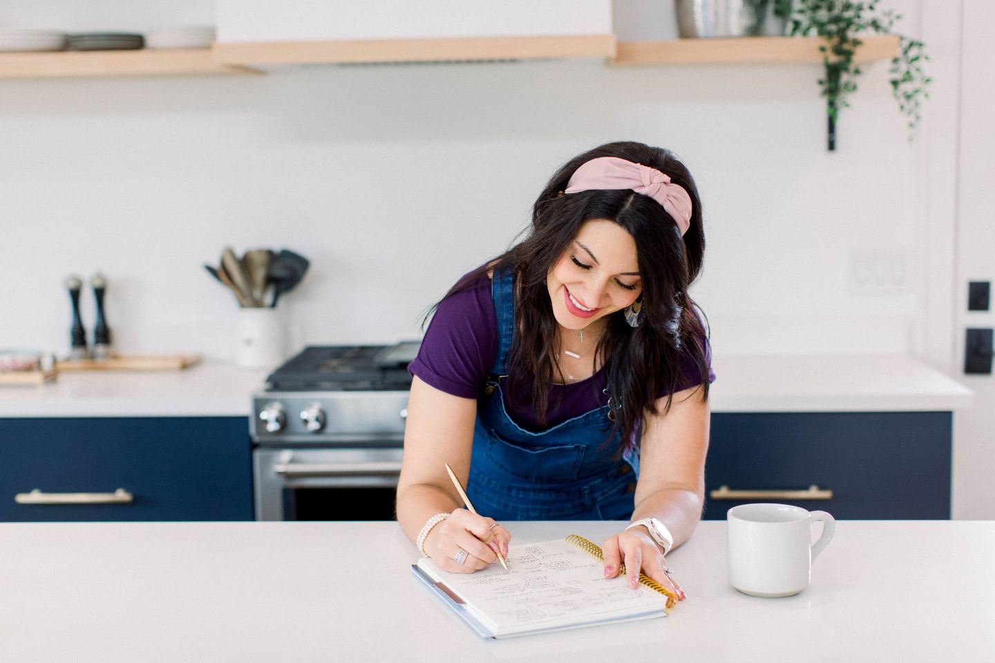 Dark haired woman working at kitchen island, writing in a notebook, coffee next to her