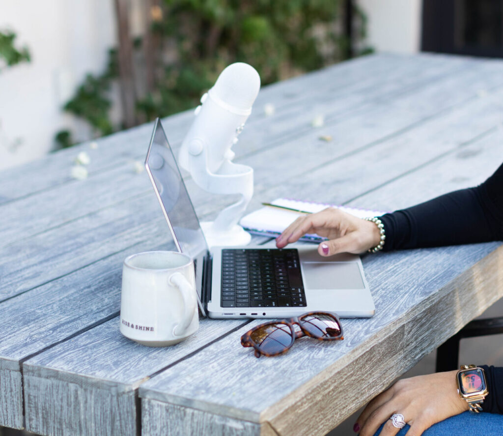 A woman's hand rest above a laptop trackpad. The laptop is sitting on a table on a wooden outdoor table. There is a podcast mic in the background and a coffee mug and pair of sunglasses in the foreground.