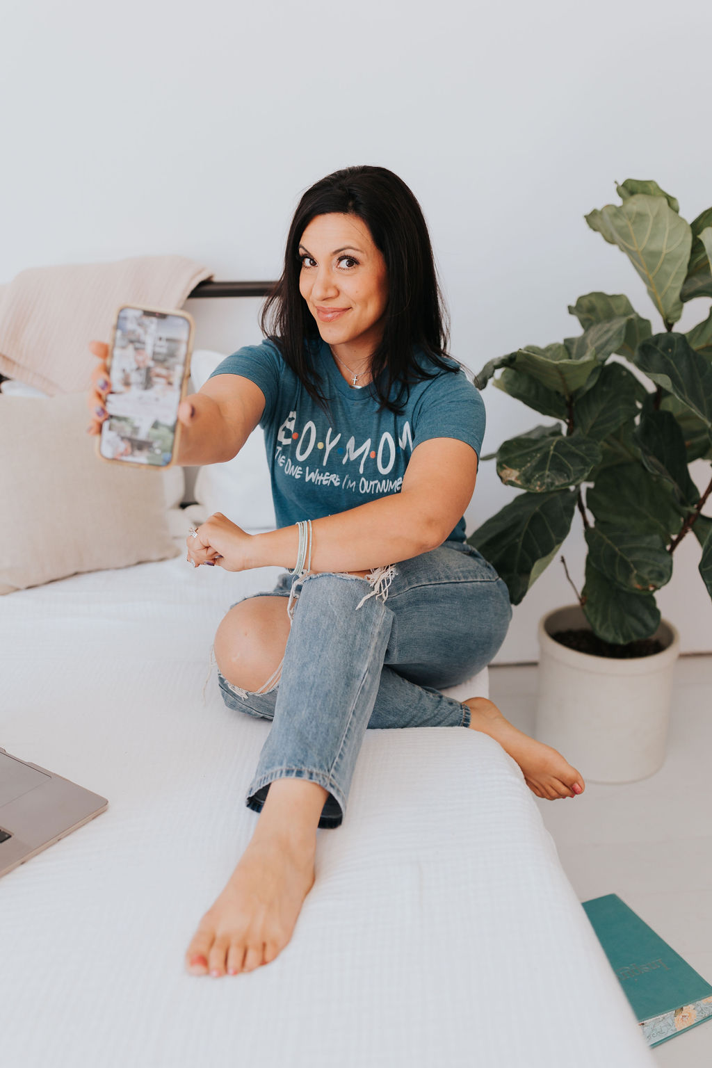 Brown haired women sitting with legs crossed, wearing jeans and teal t-shirt, holding phone out with a smile on her face, plant next to her.