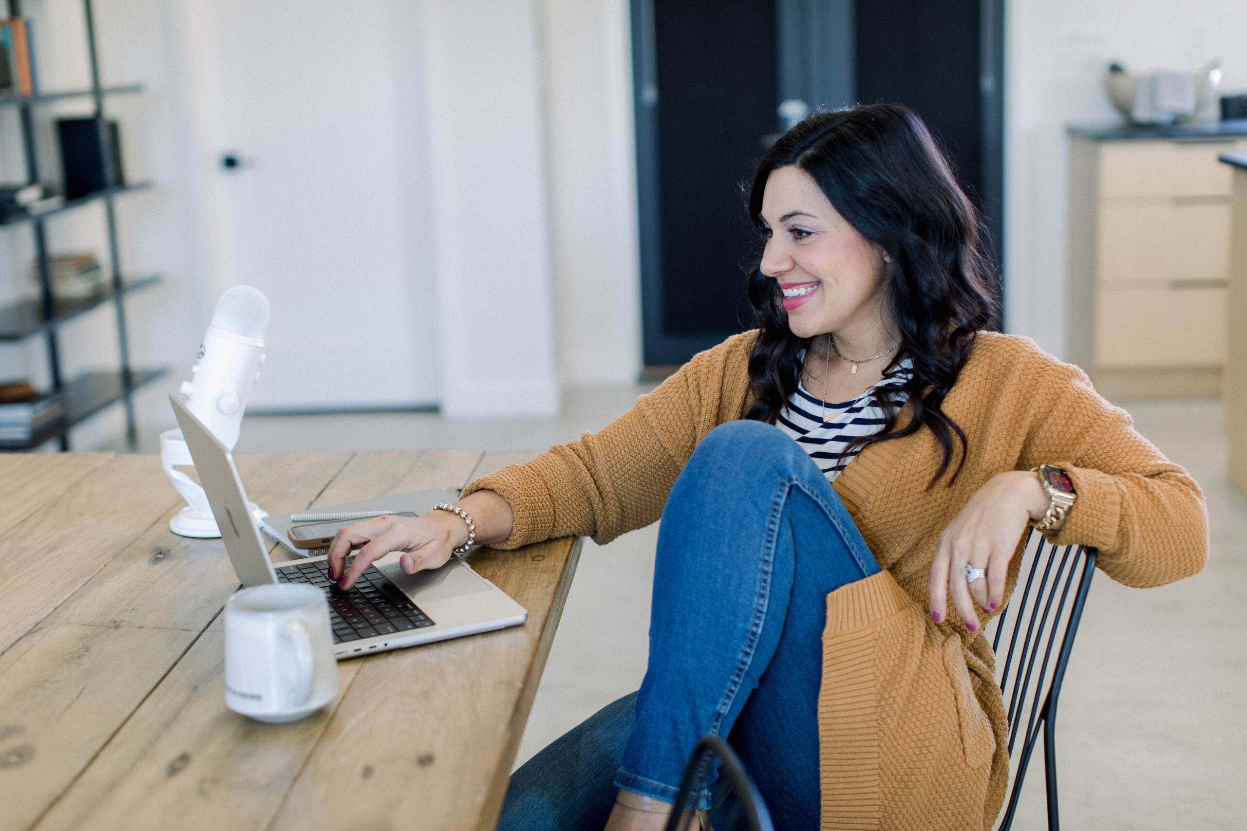 Brown haired woman sitting at desk with laptop, smiling, coffee mug, she's wearing a brown top with jeans