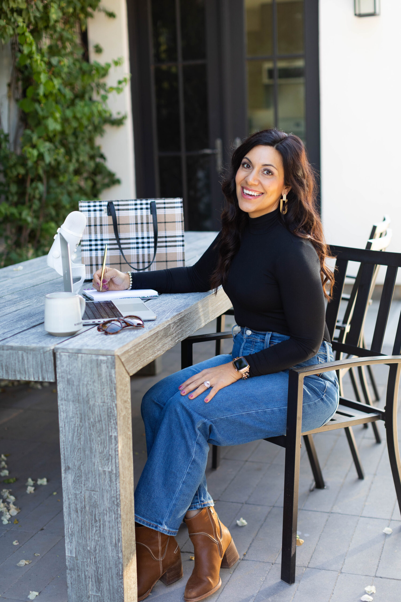Brown haired woman sitting at table outside with laptop, drink, sunglasses, purse and she's wearing a black shirt with jeans and boots.