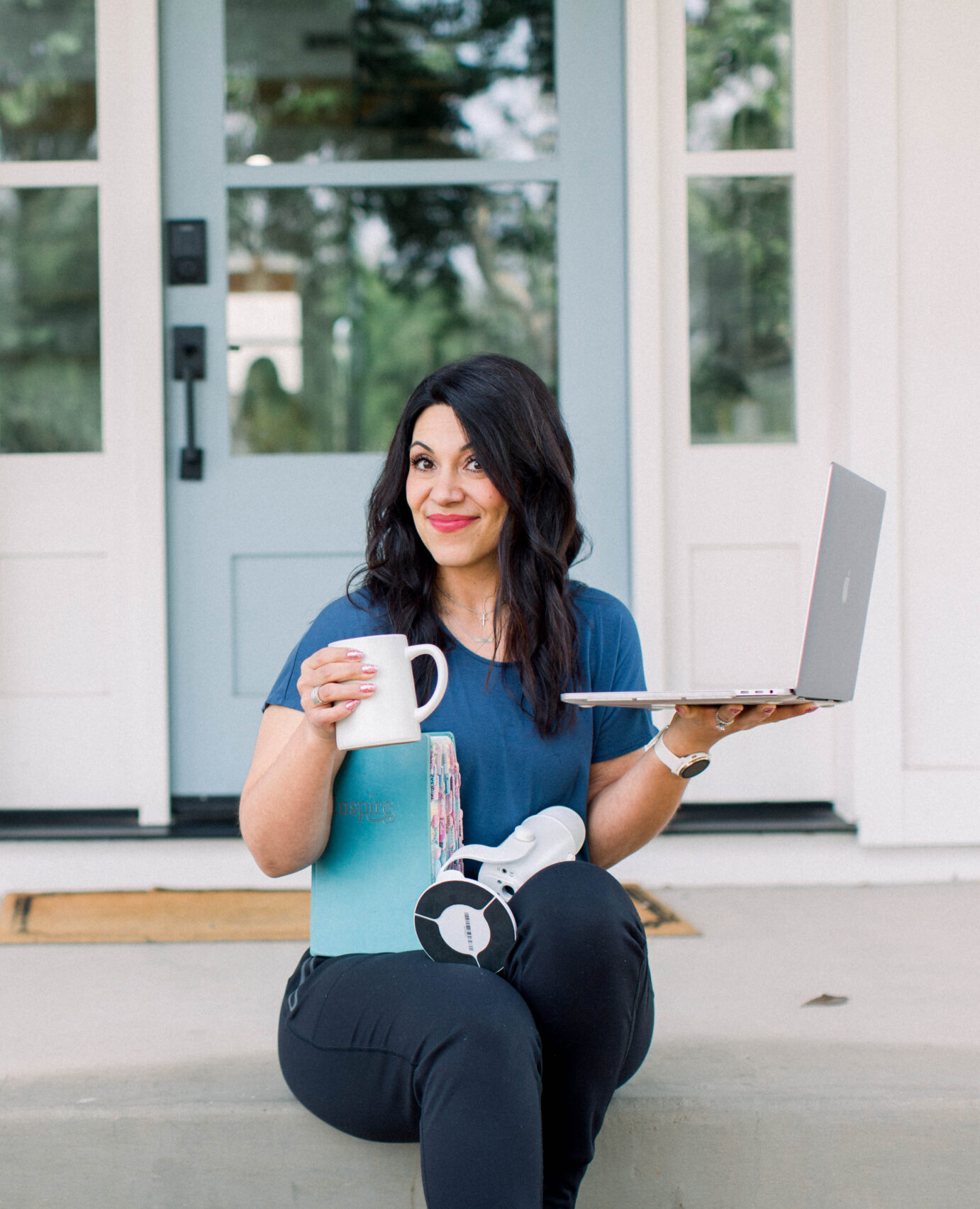 smiling brown haired woman sitting on her door step with coffee cup in hand, podcast microphone in her lap and laptop in her other hand