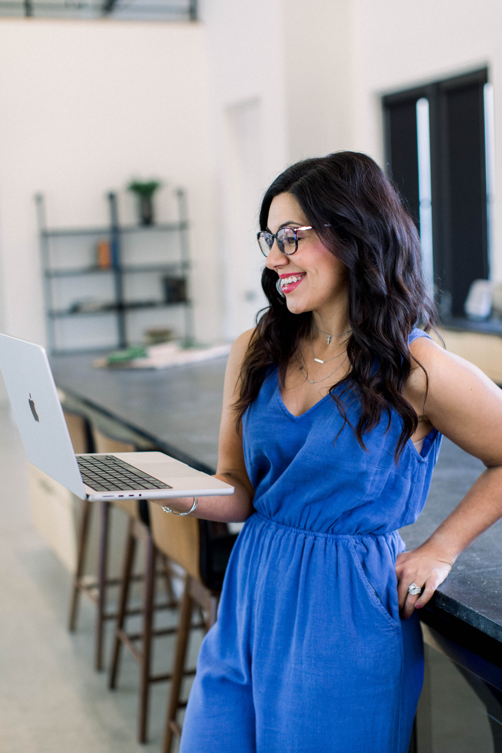 Picture of a dark haired woman wearing a blue jumpsuit. She's holding a computer leaning against a table.