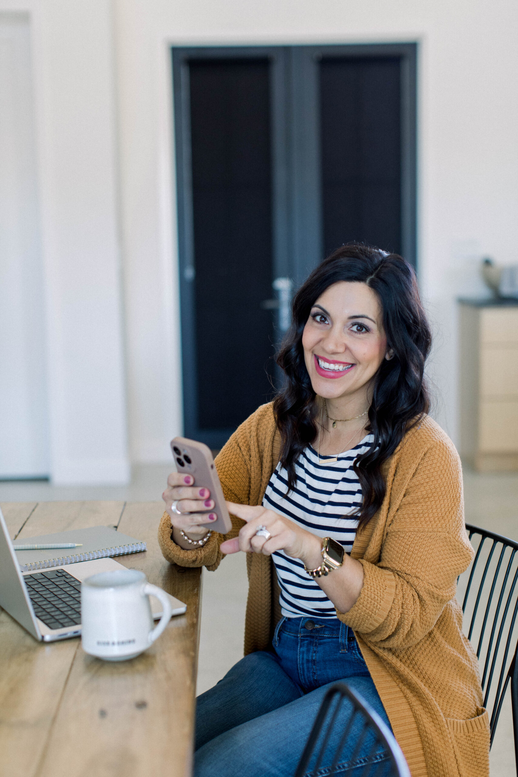 Image is a woman with dark hair sitting at a table smiling. She is holding a cell phone in her hand and there is an open laptop in front of her. 