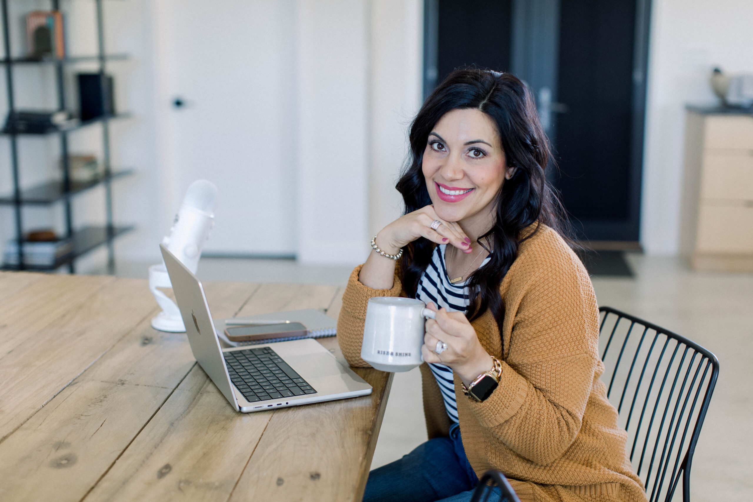 brown haired woman smiling as she works at her desk with her laptop, coffee, and podcast microphone