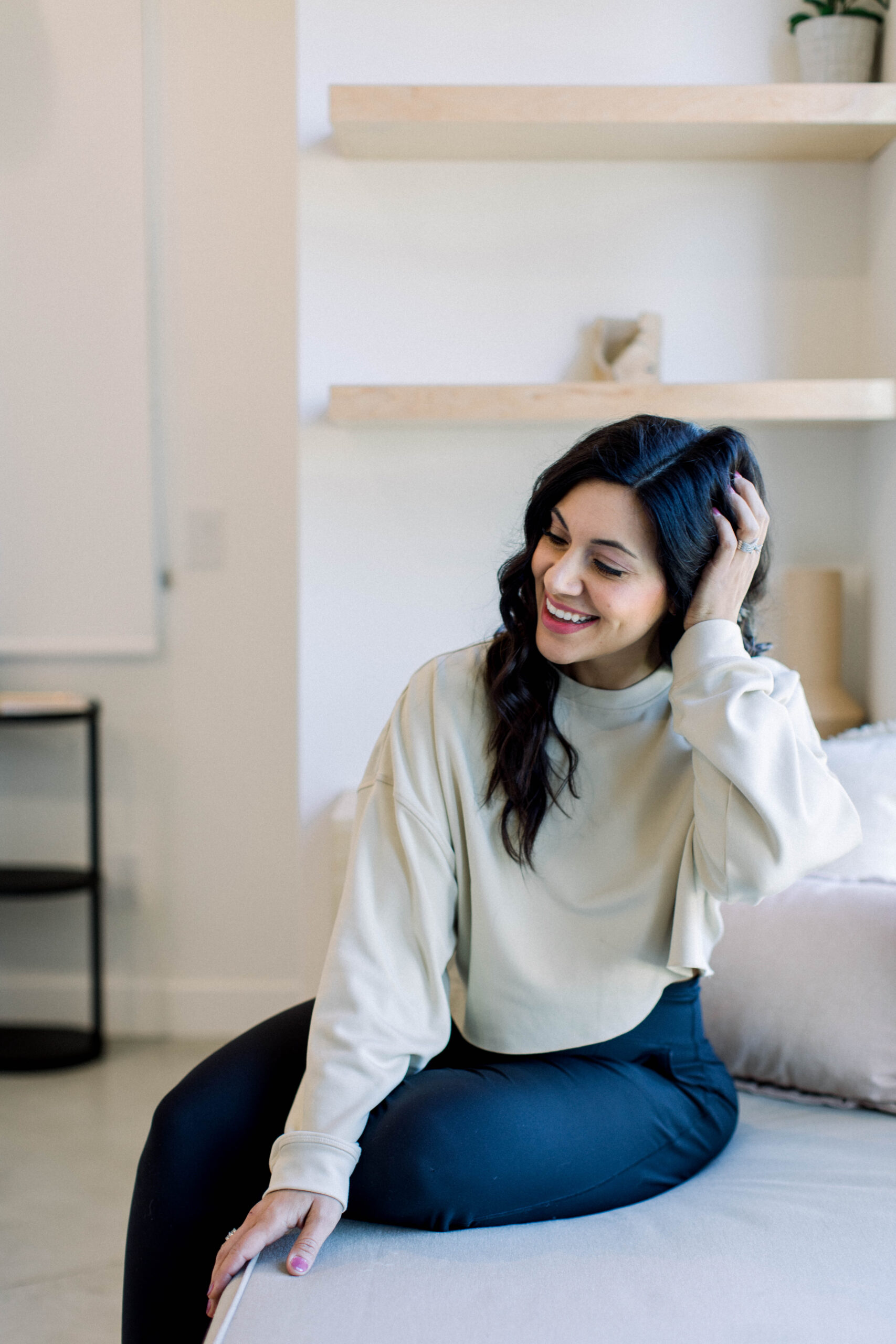 Image is of a woman with dark hair smiling while sitting on her bed