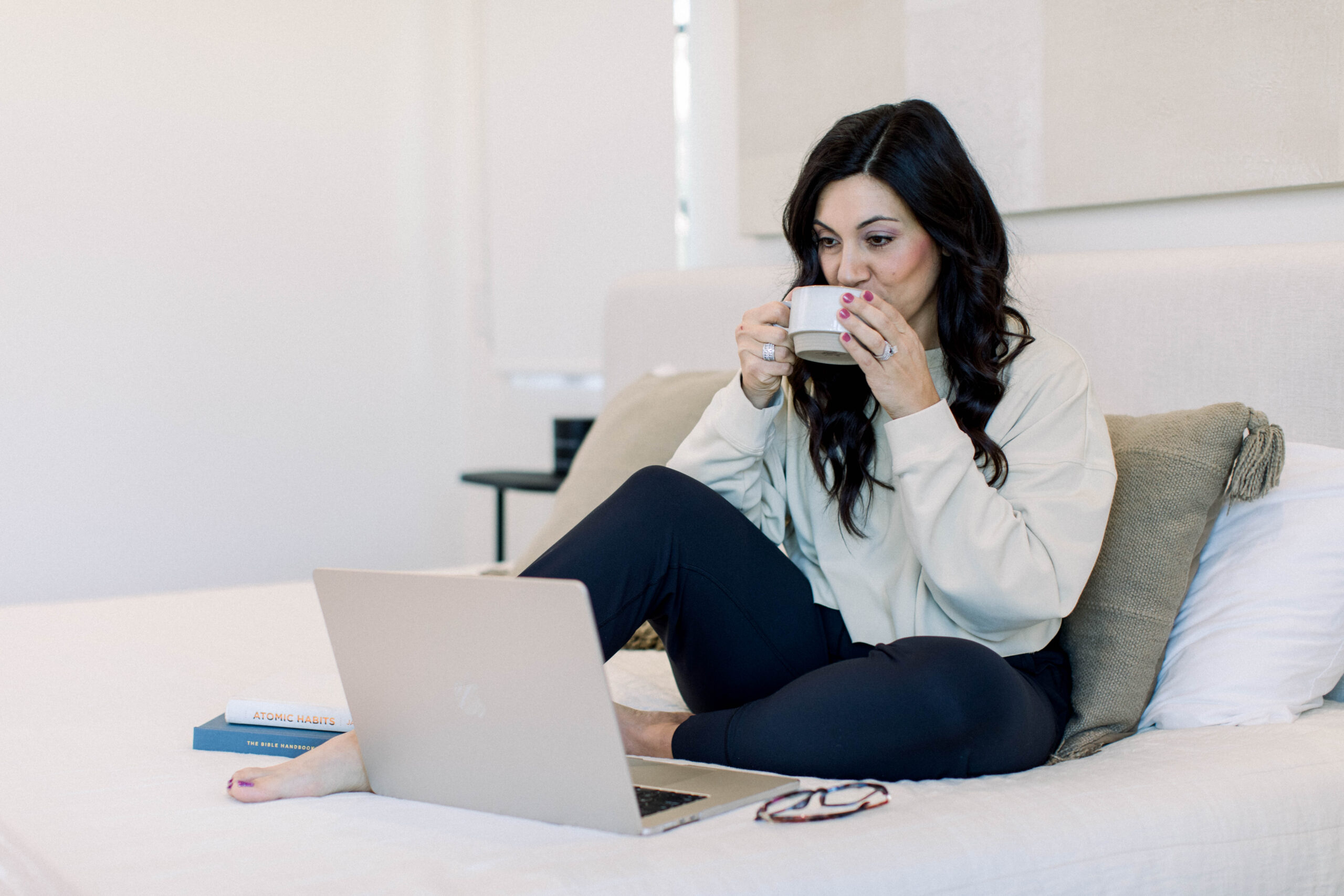 Image is a woman with dark hair drinking coffee on her bed while looking at her computer.