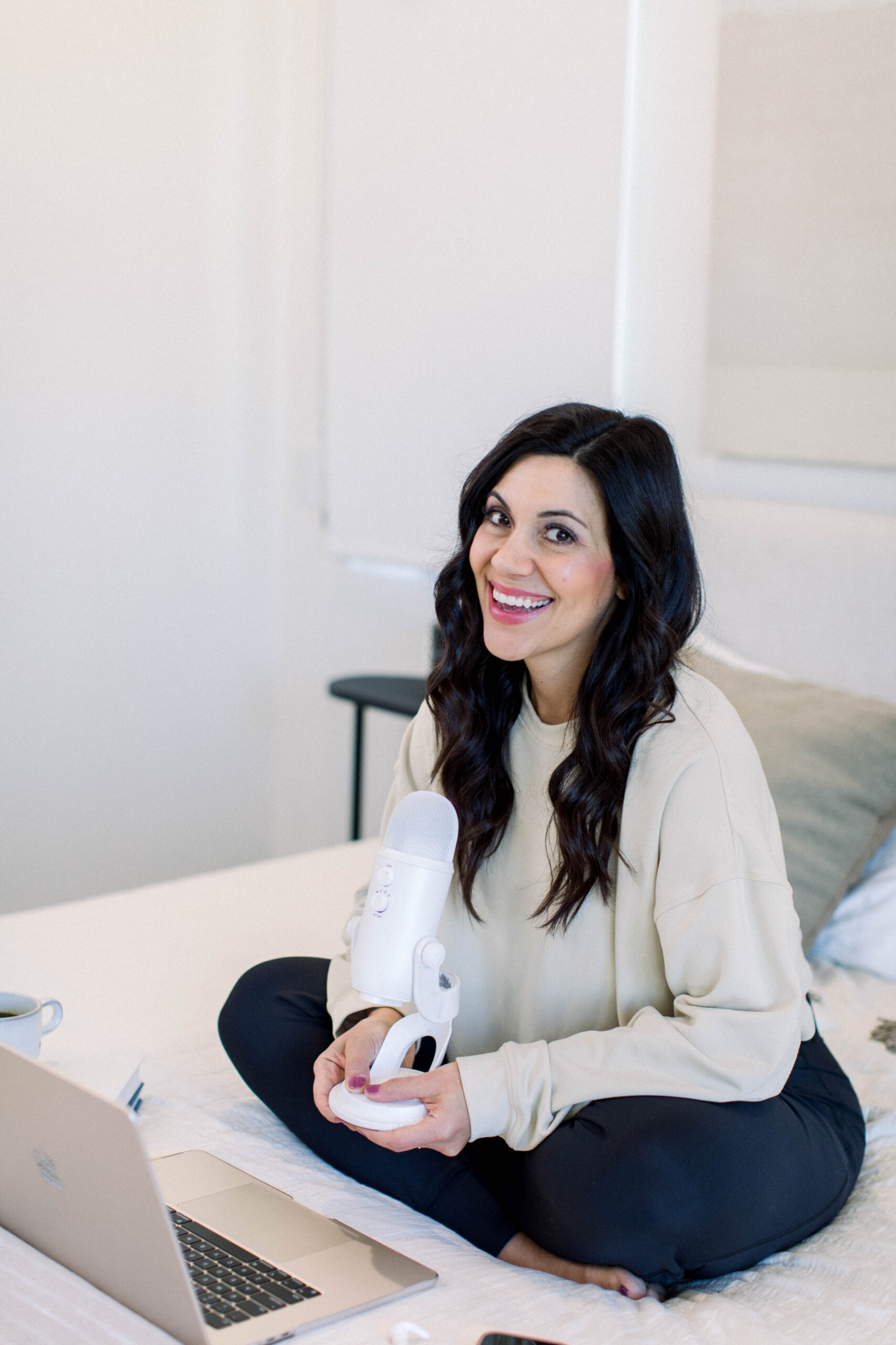 Image is of a woman with dark hair sitting on a bed with a microphone in hand. 