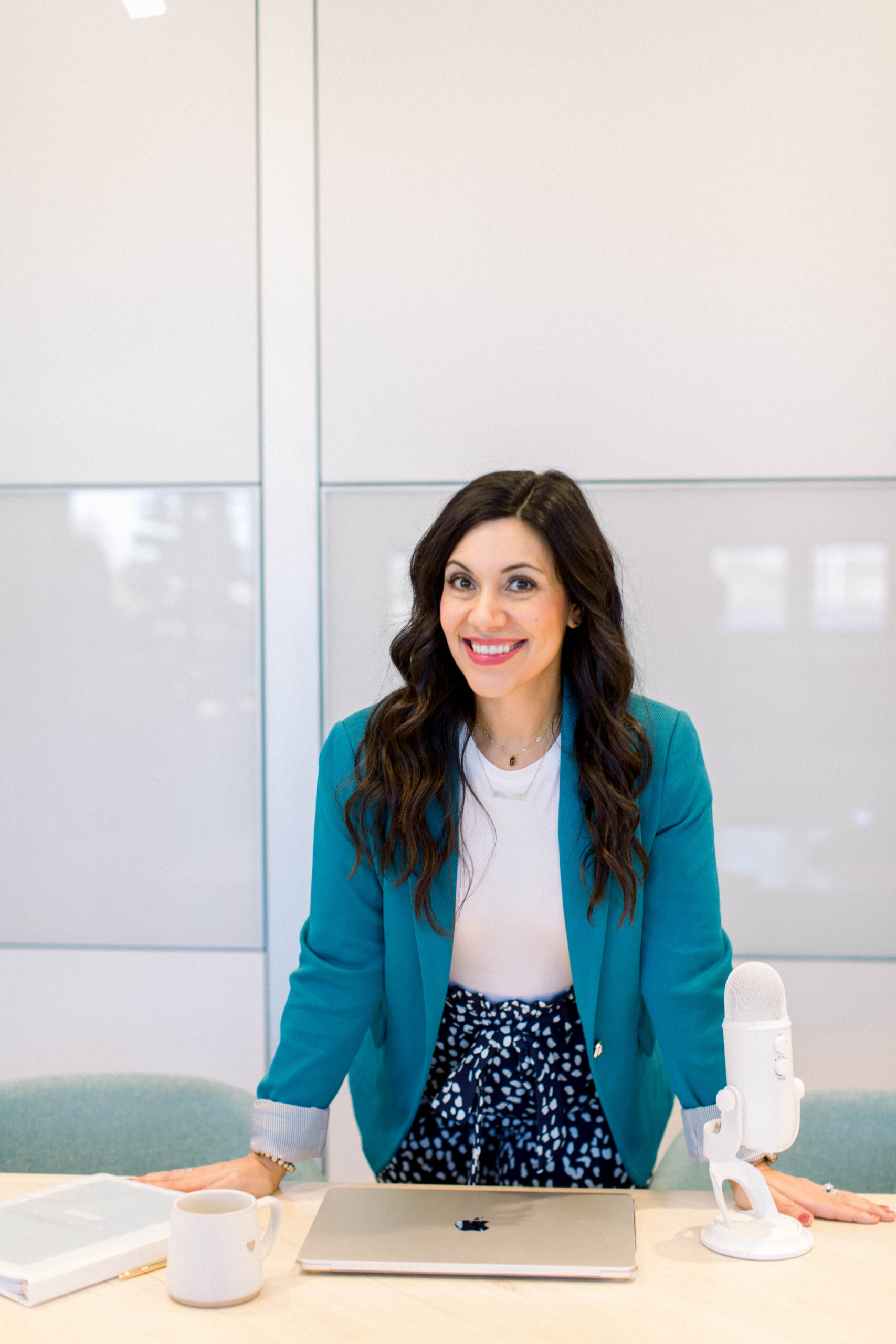 Image is of a smiling woman with dark hair wearing a business suit leaning against a table.