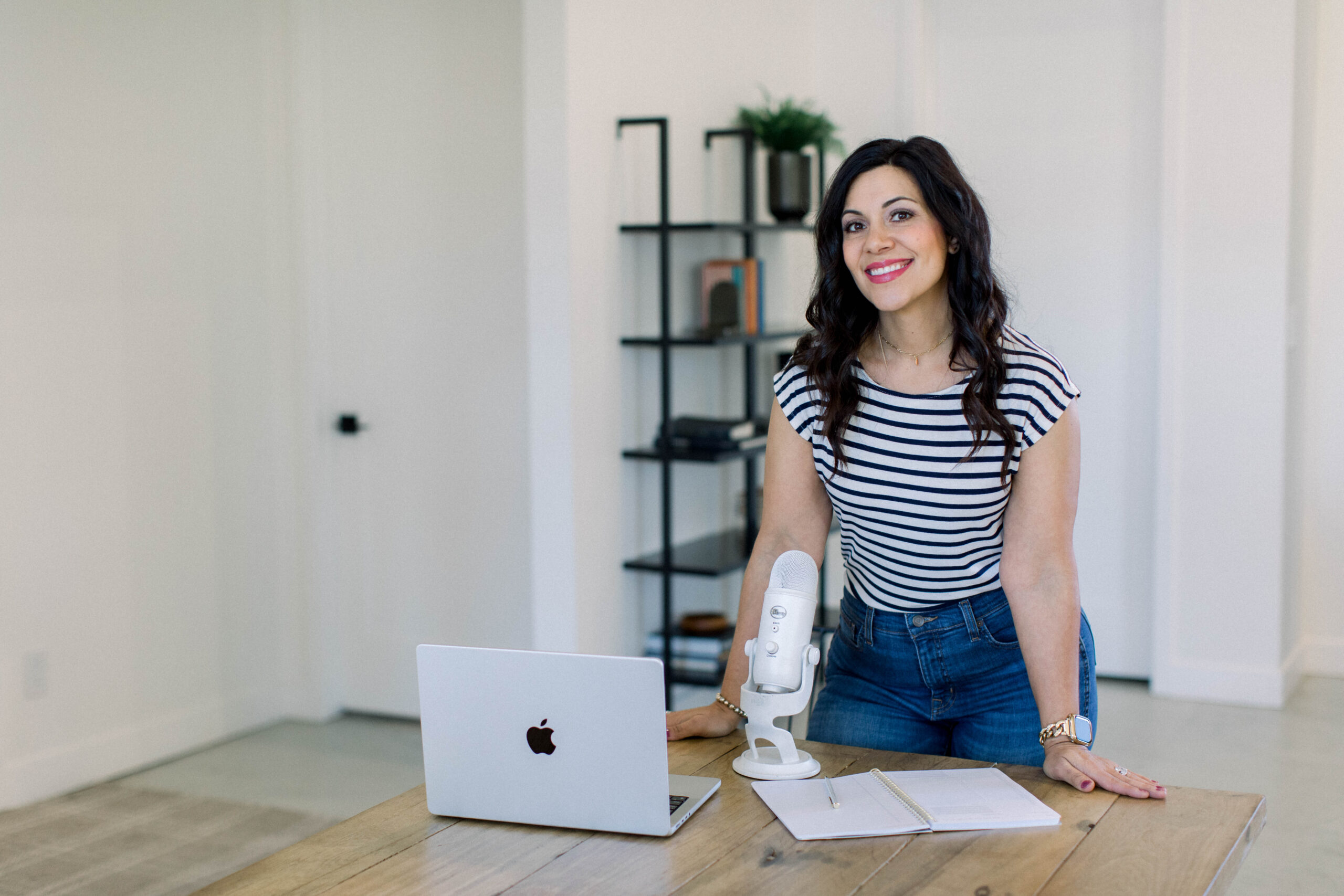 smiling brown haired woman standing at her table with her white podcast microphone and laptop.